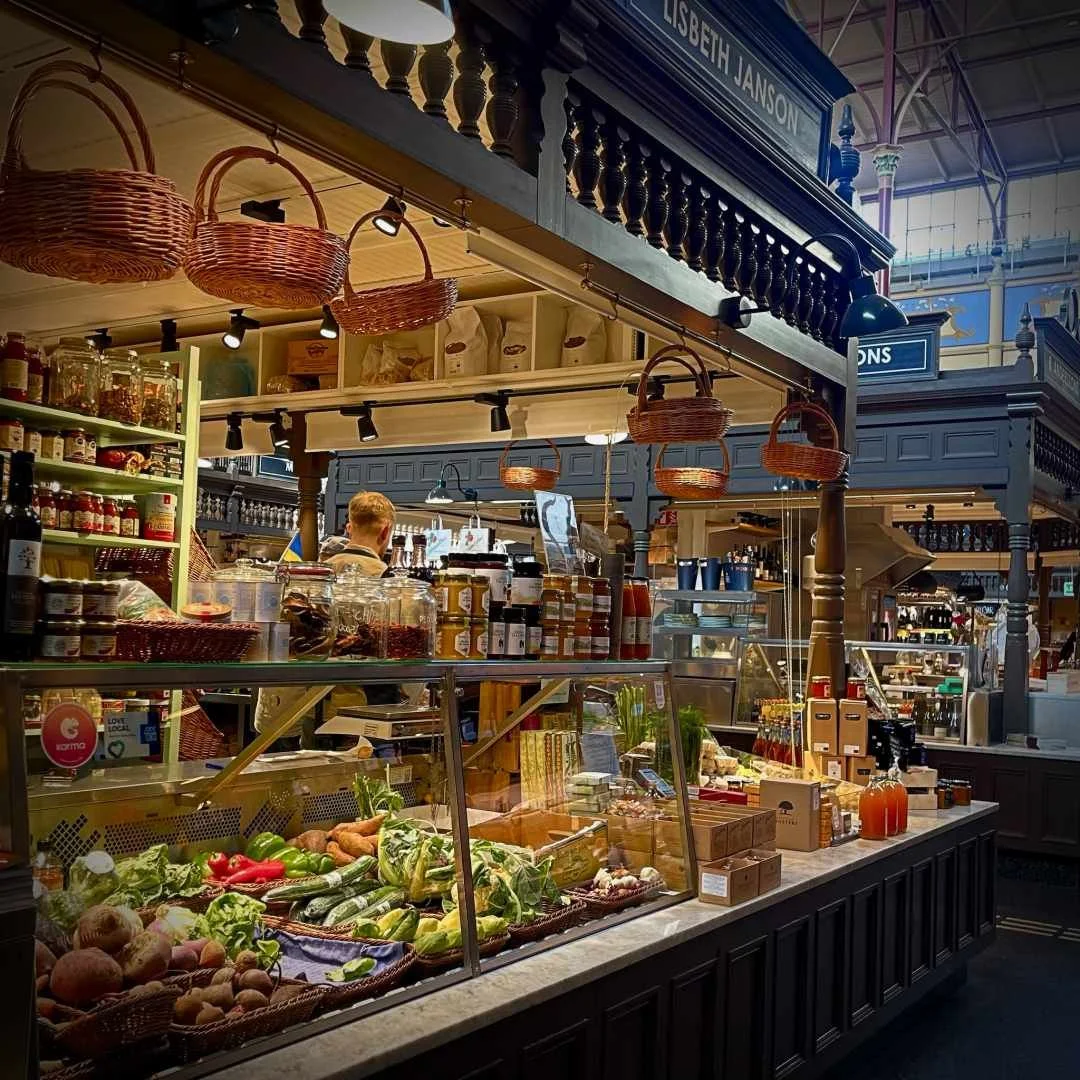 Der Verkaufsstand von „Lisbeth Janson“ in der Östermalms Saluhall. Unter hängenden Weidenkörben wird in einer Glasvitrine frisches Gemüse präsentiert, auf der Theke daneben stehen Gläser mit Feinkost und Säfte.