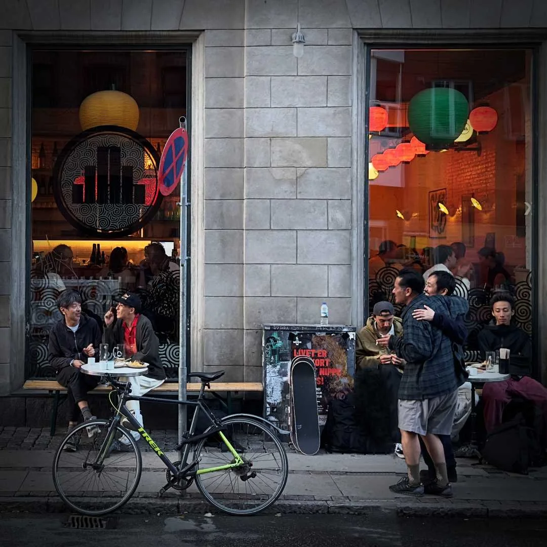 Aussenansicht eines japanischen Izakaya-Restaurants mit dem Namen Jah. Mehrere Personen sitzen und stehen draußen an kleinen Tischen. Japanische Laternen sind im Innenraum sichtbar. Ein schwarzes Fahrrad steht davor.