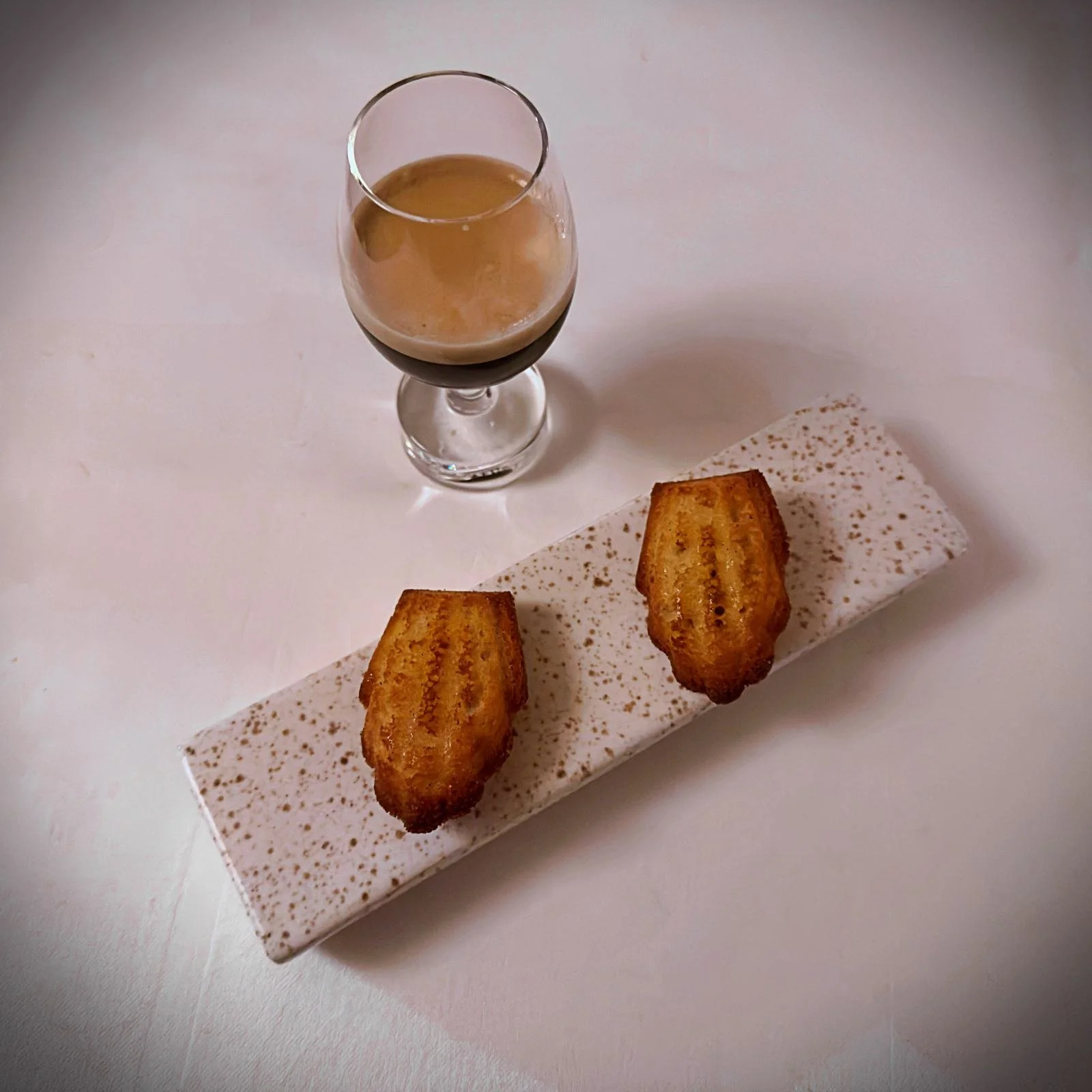 Zwei goldbraune Madeleines liegen auf einer länglichen, gesprenkelten Platte. Daneben steht ein Glas mit Espresso. Es ist ein Dessertgang im Restaurant R21 in Amsterdam auf hellem Untergrund.