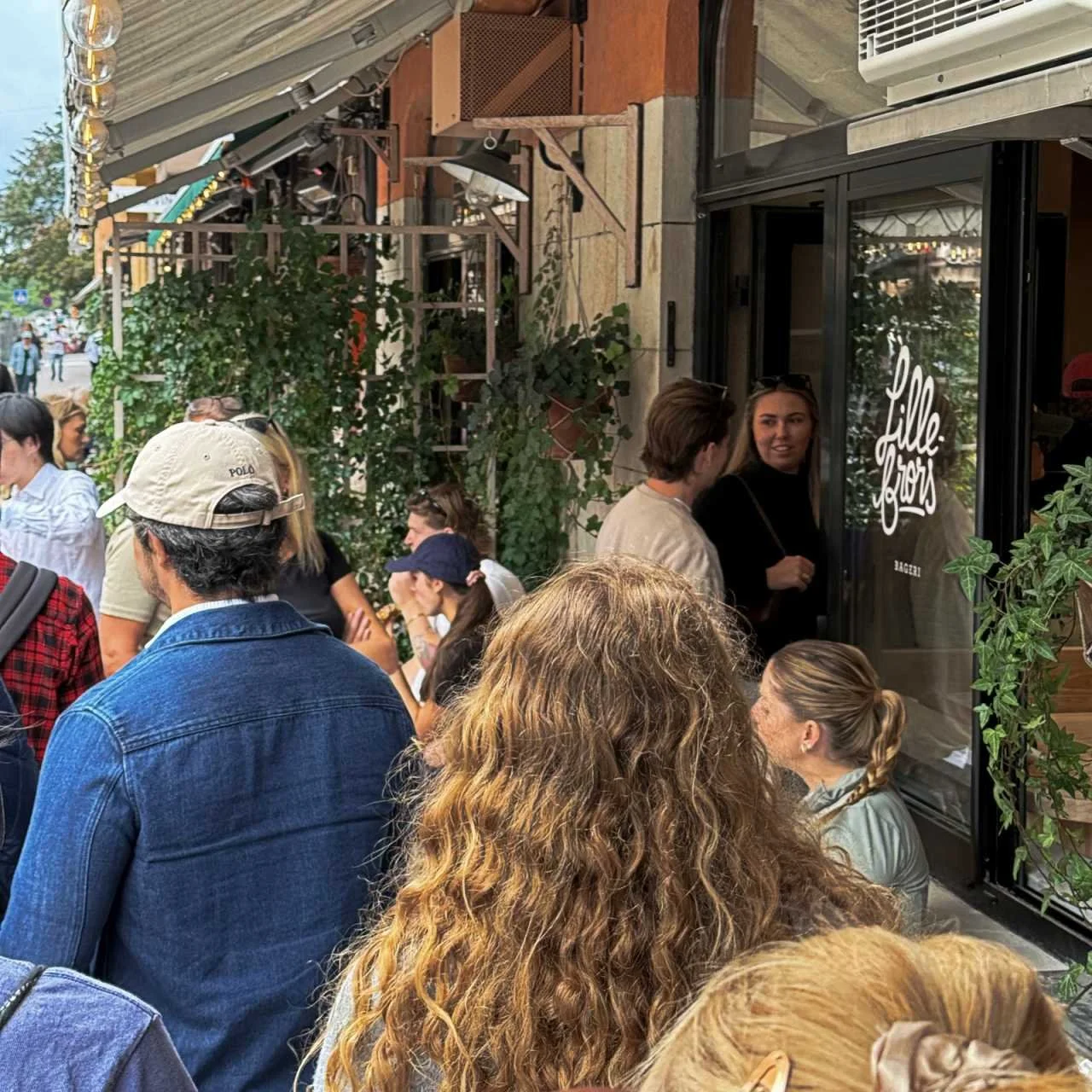Eine Gruppe von Menschen steht auf dem Gehweg vor der Eingangstür der Bäckerei Lillebrors Bageri an. An der Hausfassade hängen Rankpflanzen und eine Markise.