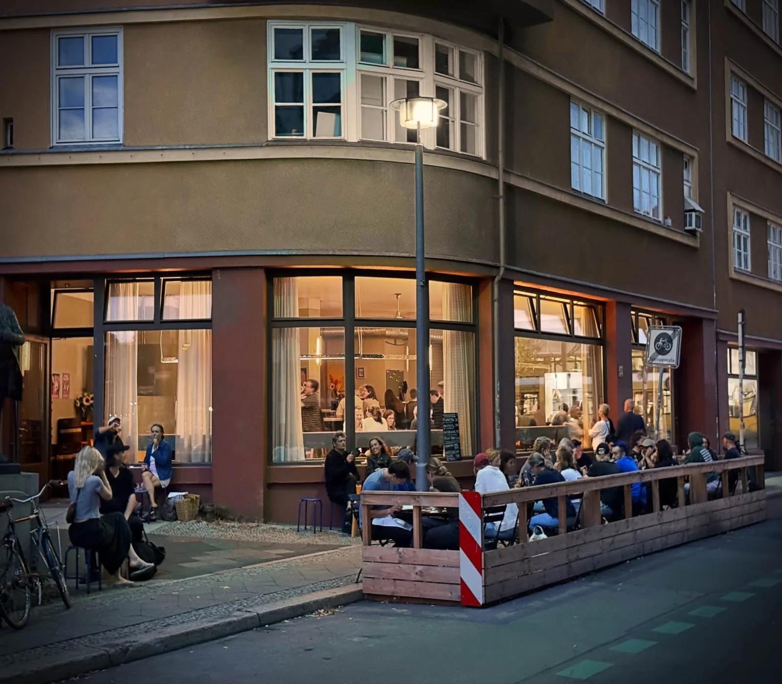 Exterior view of Restaurant Trio on a building corner with illuminated windows. A wooden-fenced outdoor seating area with guests is set up on the street.