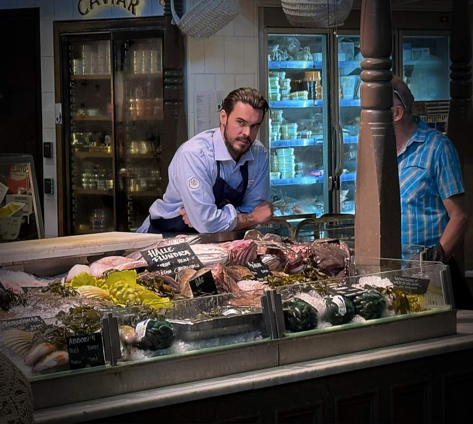 A fishmonger with a beard and tattoos leans behind his counter in Östermalms Saluhall, wearing a light blue shirt and dark apron. Various fish specialties such as halibut (Hälleflundra) and perch (Abborre) are displayed on ice in front of him.