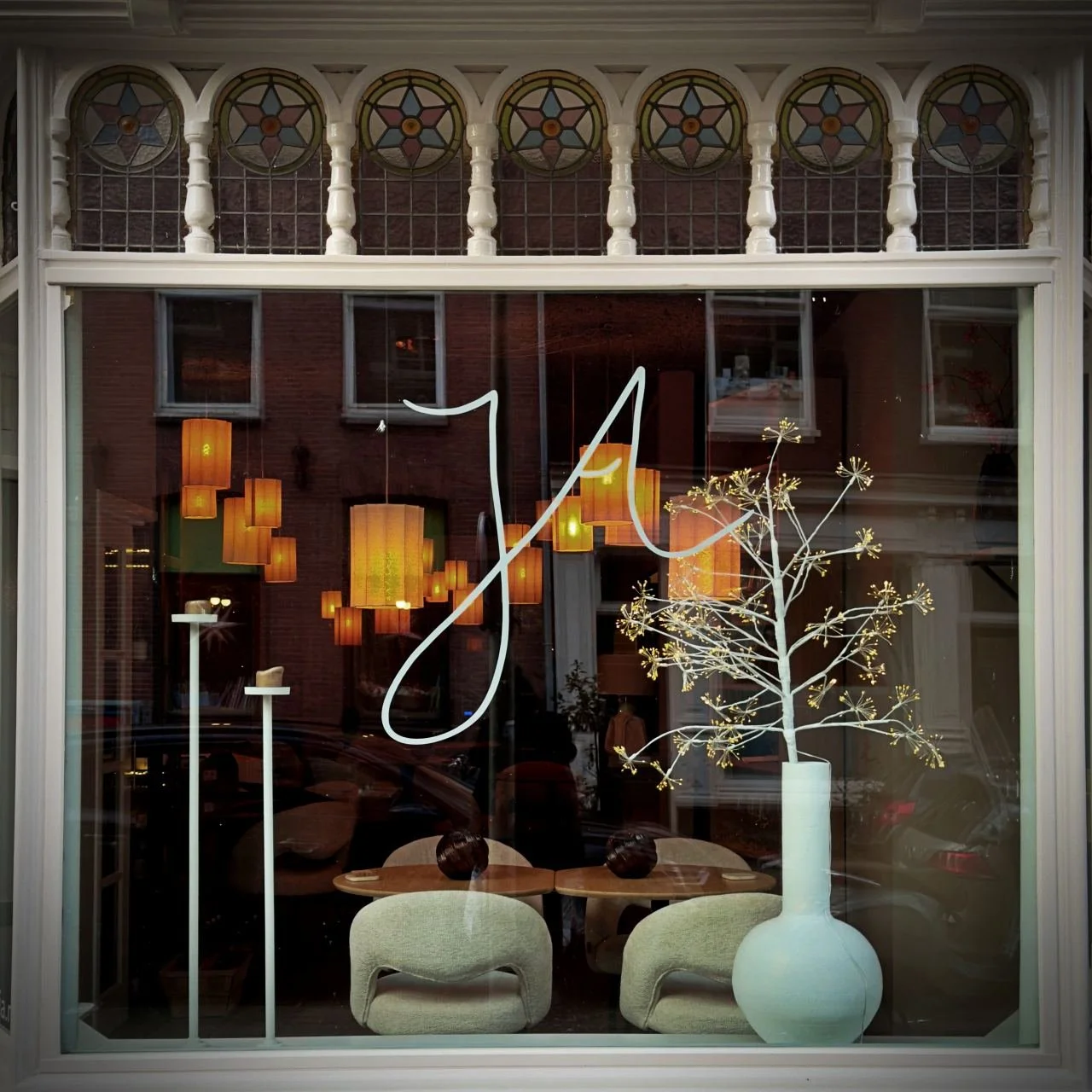 View through the window of Restaurant Jā in Amsterdam with floral vase, pastel lounge chairs, pendant lights and large white logo on the glass pane.