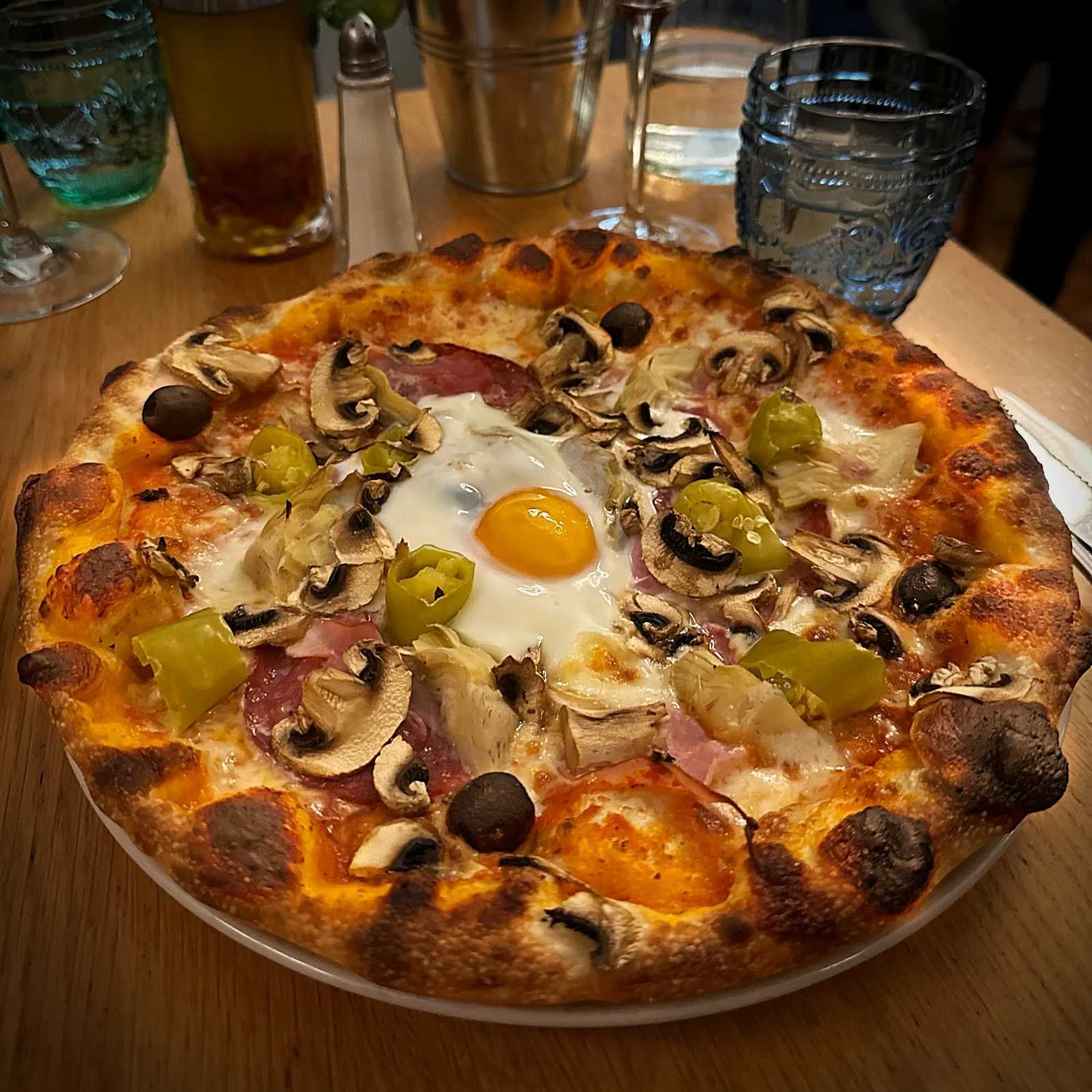 Pizza at Il Piccolo Principe restaurant in Munich with mushrooms, olives, peppers, artichokes, and a fried egg in the center on a wooden table.