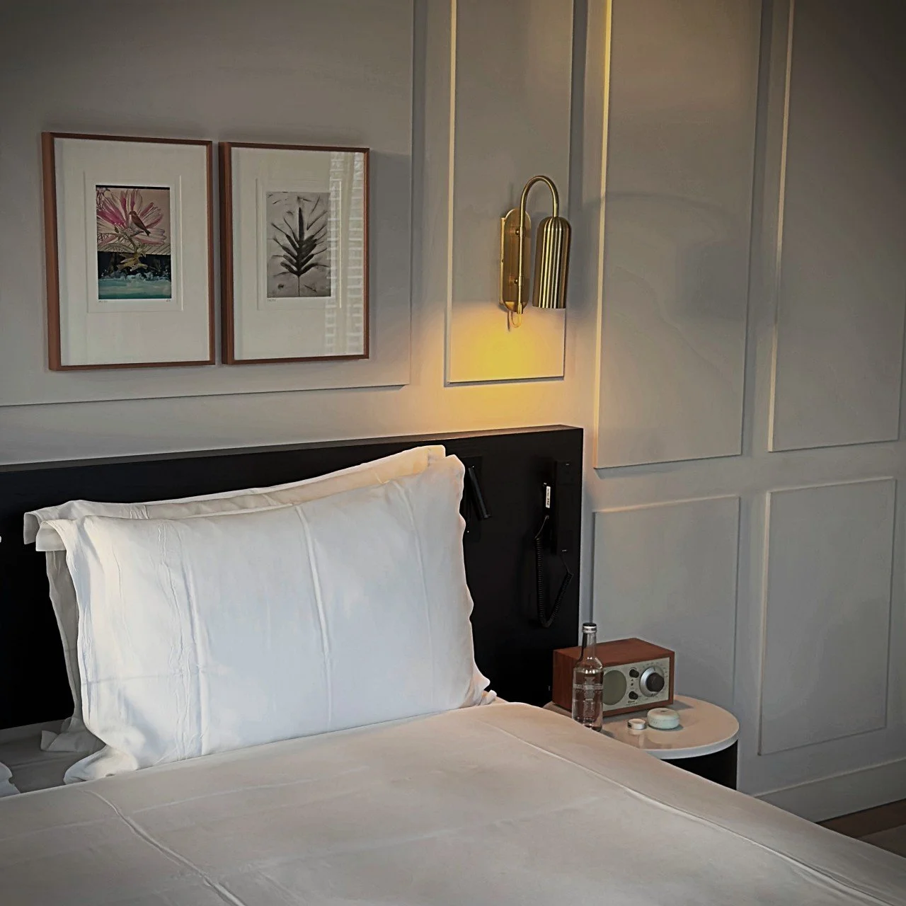 Nahaufnahme eines Hotelbettes mit weißer Bettwäsche, zwei gerahmten Bildern an der Wand und einer messingfarbenen Wandleuchte.