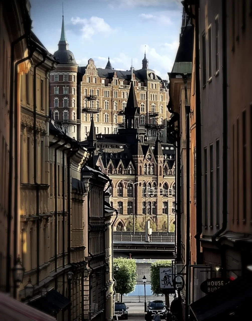 View through a narrow street in Gamla Stan toward historic rows of buildings with detailed facades. Brick architecture and towers of central Stockholm in the background.