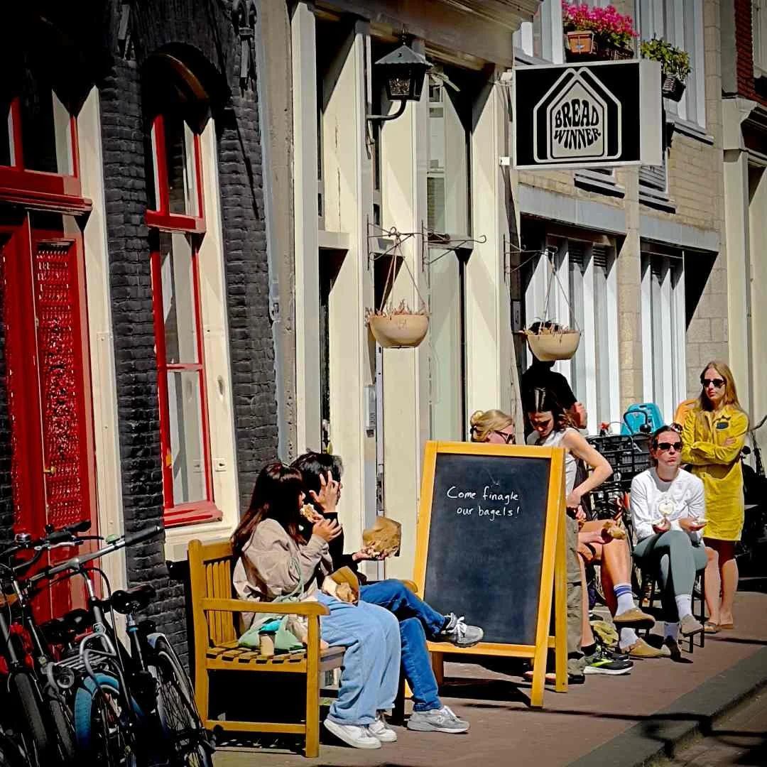 Menschen genießen Bagels vor dem Breadwinner Café -Jordaan in Amsterdam. Ein Schild mit der Aufschrift „Come finagle our bagels!“ steht auf dem Bürgersteig.