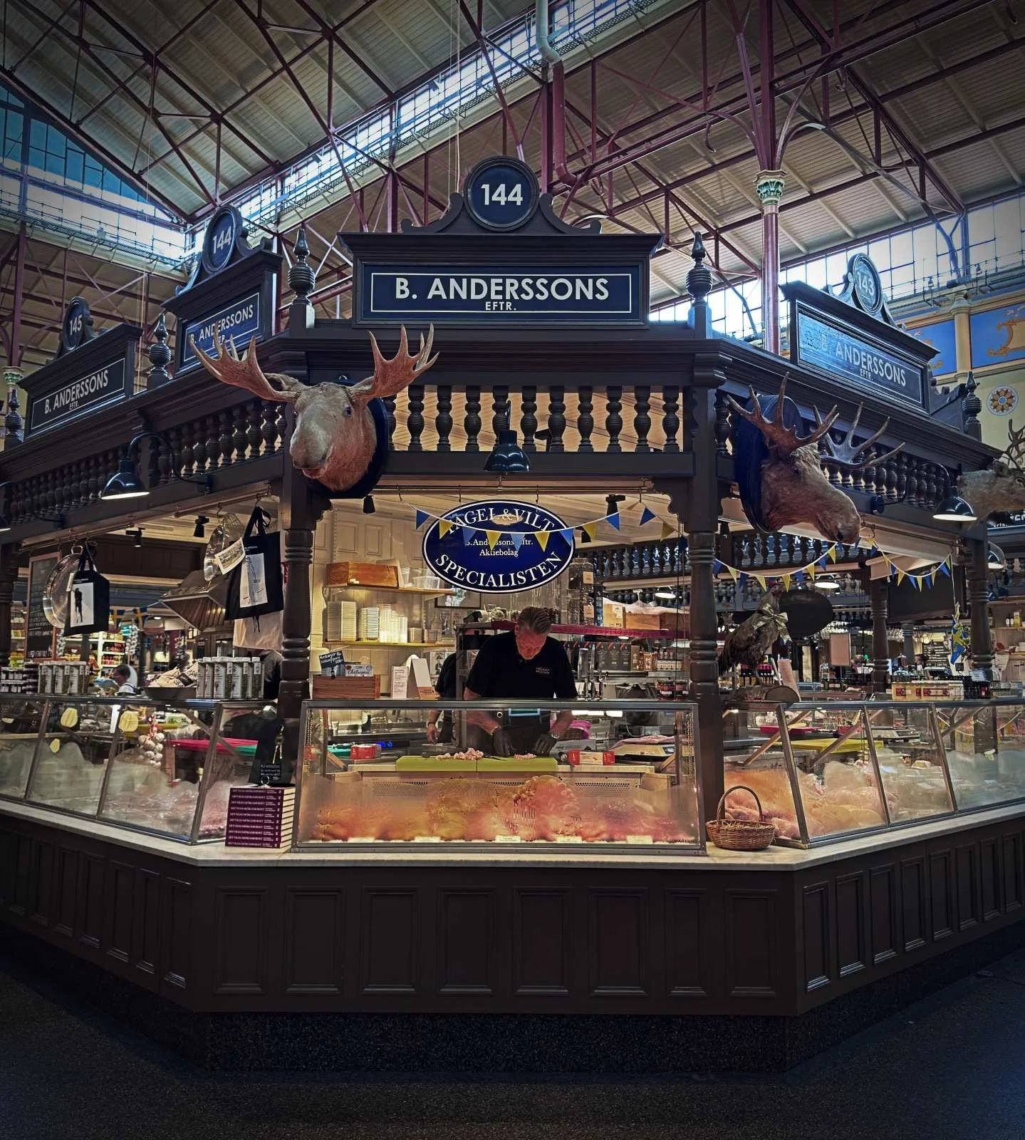 The "B. Anderssons" stall in Östermalms Saluhall. Two mounted elk heads hang from the wooden structure above the glass counter, where game specialties are displayed.