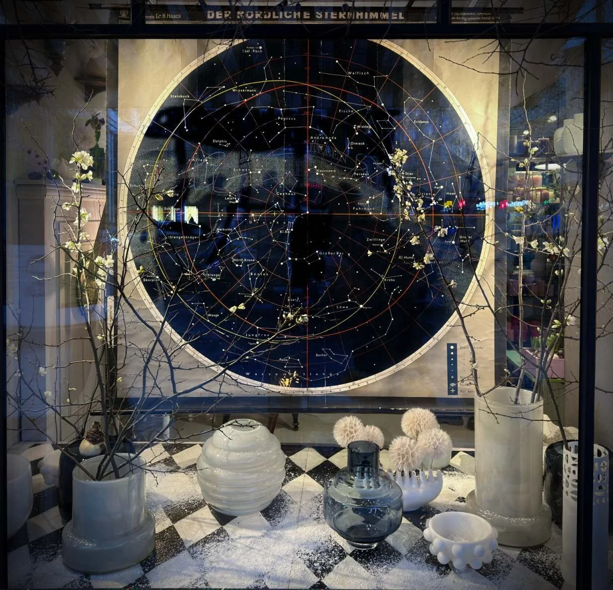 The storefront of the shop "Blumen die Leben" in Munich features a vintage school poster of the starry sky as a backdrop. In front of it, various glass vases and decorative objects are arranged on a black and white checkered floor.
