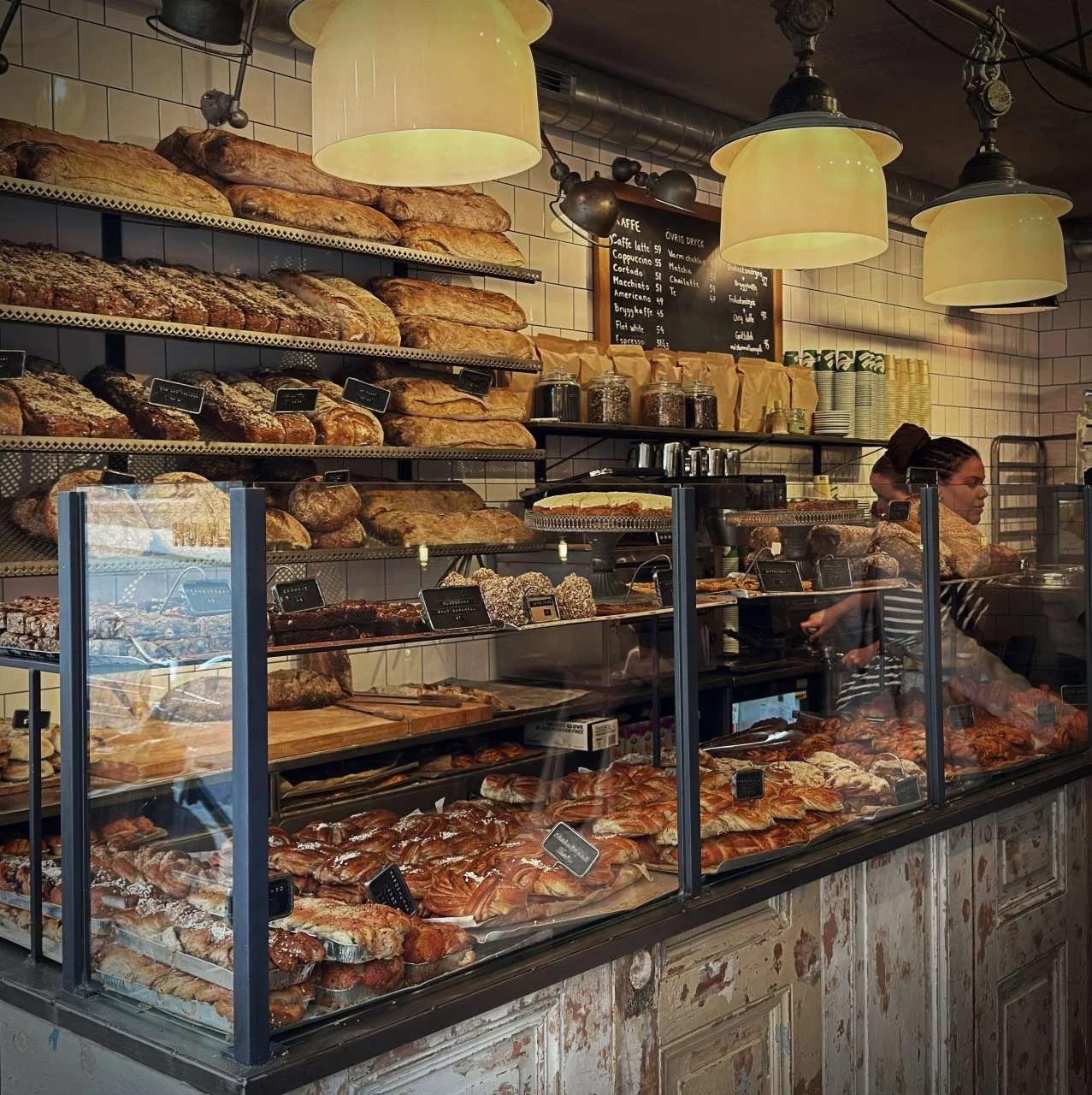 Blick in die Bäckerei: Hinter der Theke stapeln sich frische Brote in den Regalen. Vorne in der Vitrine liegen Zimtschnecken und Kuchen bereit. Im Hintergrund steht ein alter, weißer Kachelofen.
