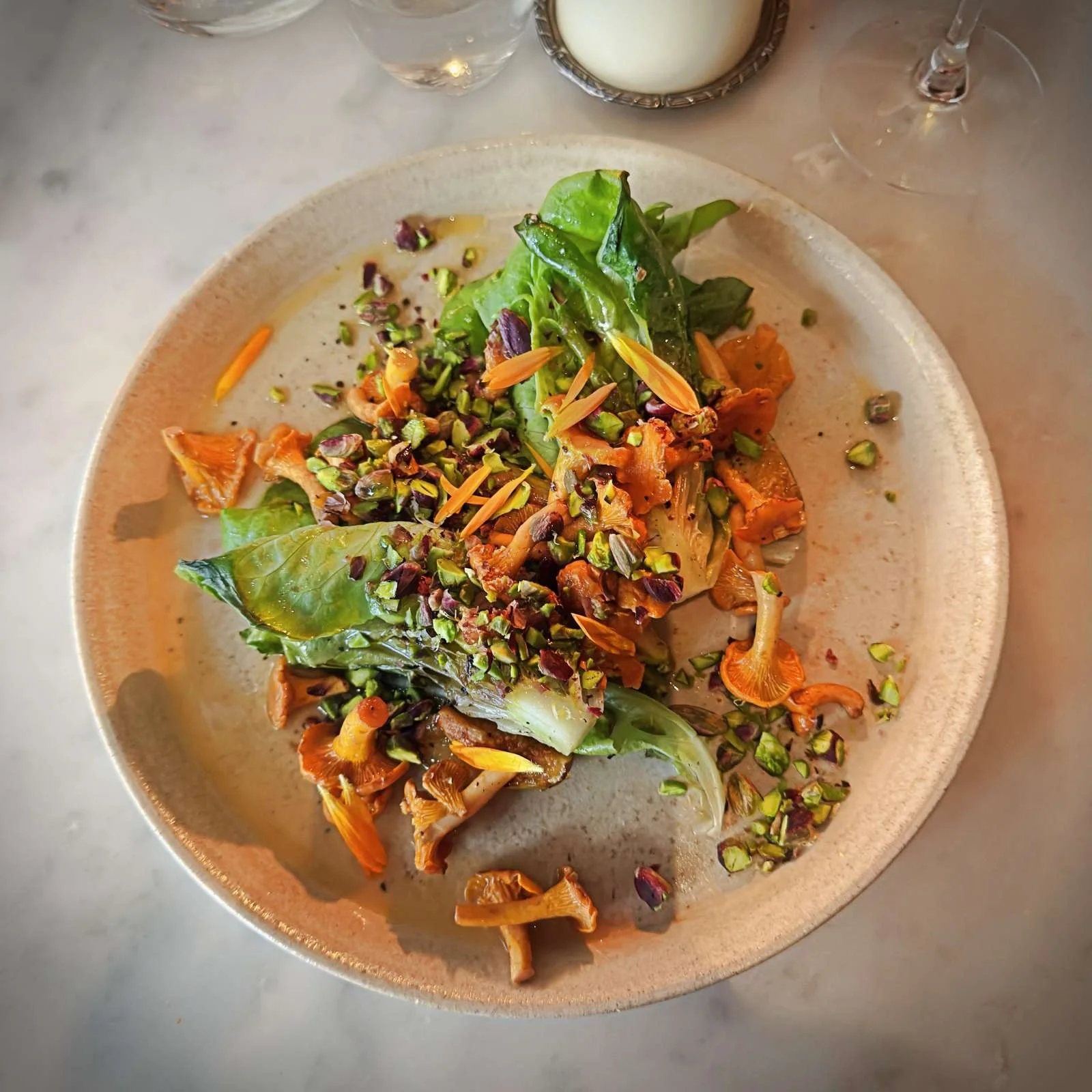Gegrillter Salat serviert mit Pfifferlingen und gehackten Pistazien auf einem beigefarbenen Teller auf einem Marmortisch.