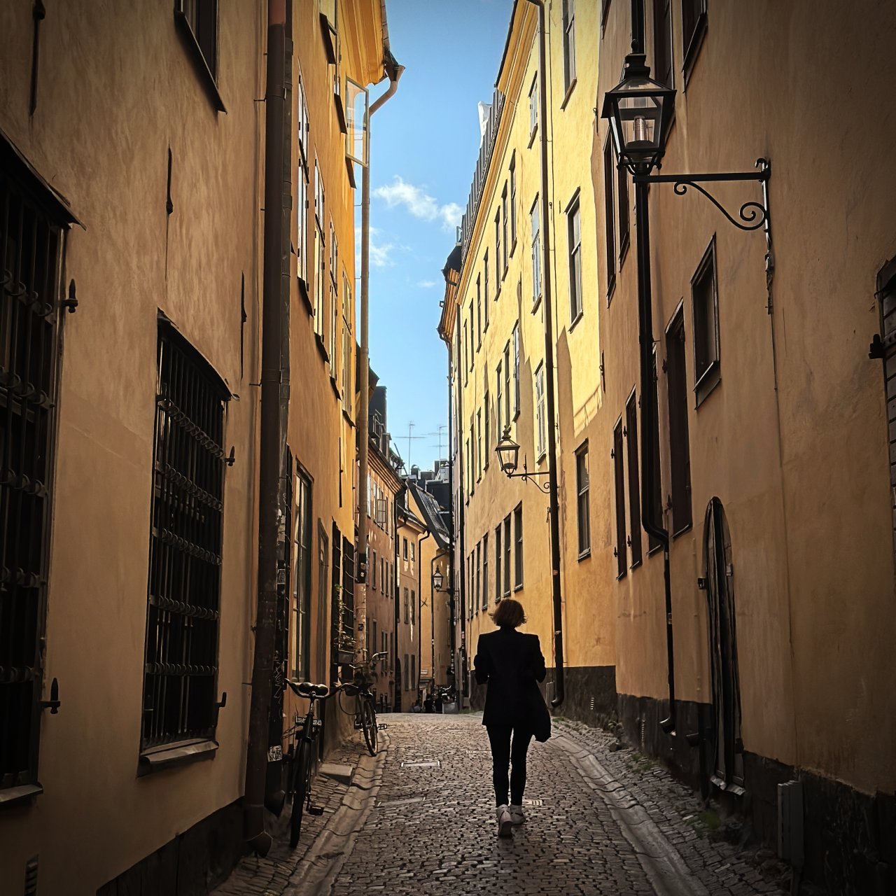 Schmale Kopfsteinpflasterstraße in Gamla Stan, Stockholm. Gelbe Hausfassaden, historische Straßenlaternen und Fahrräder am Rand. Eine Person geht mittig durch die Gasse.