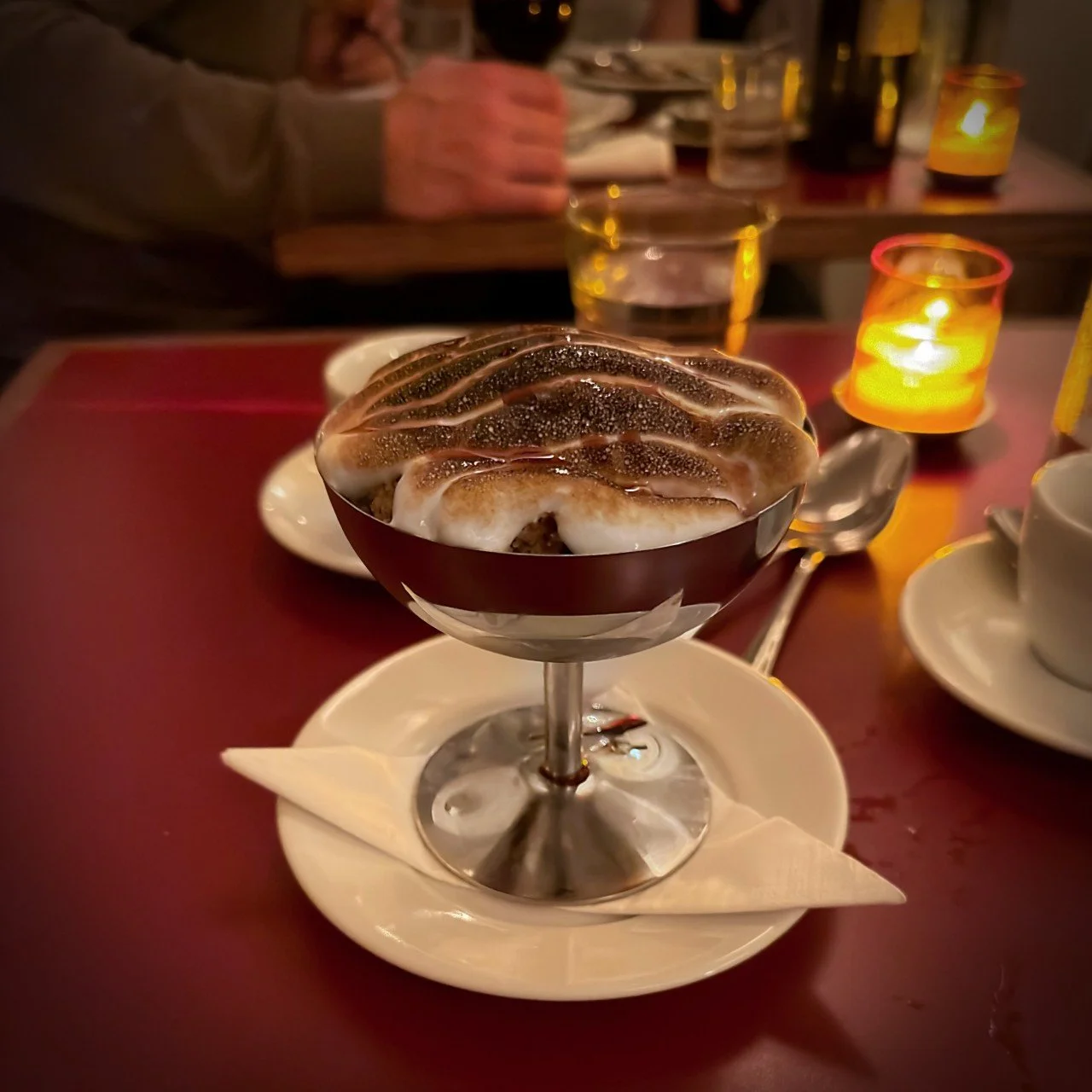 Flambéed meringue crowns the cherry ice cream coupe in a silver cup. In the background, a tea light glows on the table.