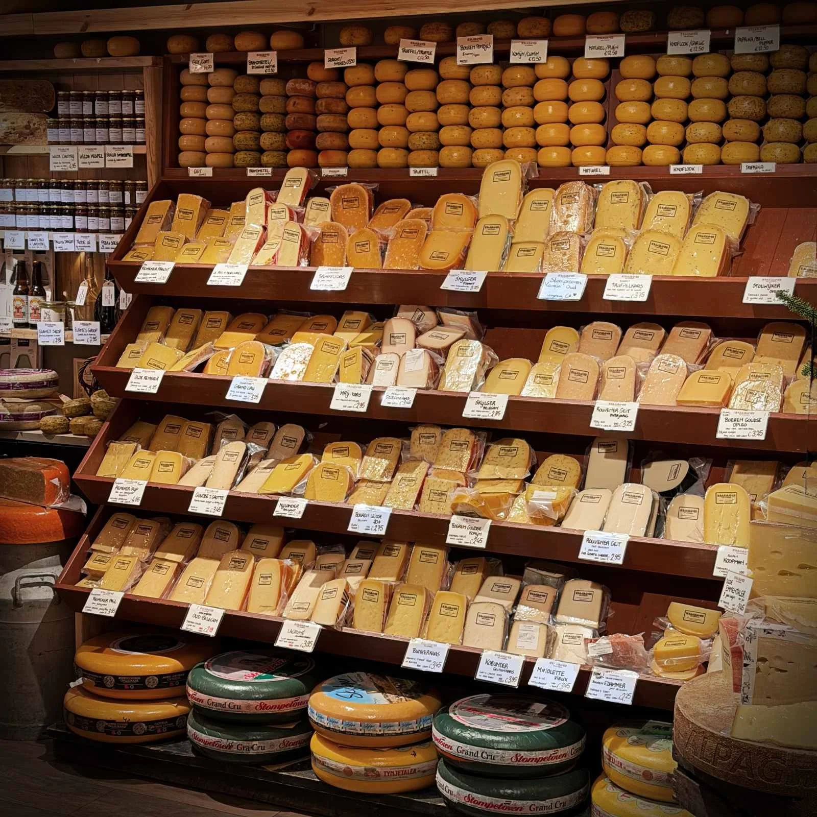 At the De Kaaskamer shop in Amsterdam, tall wooden shelves are filled with various types of cheese. Packed cheese portions with price tags and whole wheels are displayed in rows on the shelves.