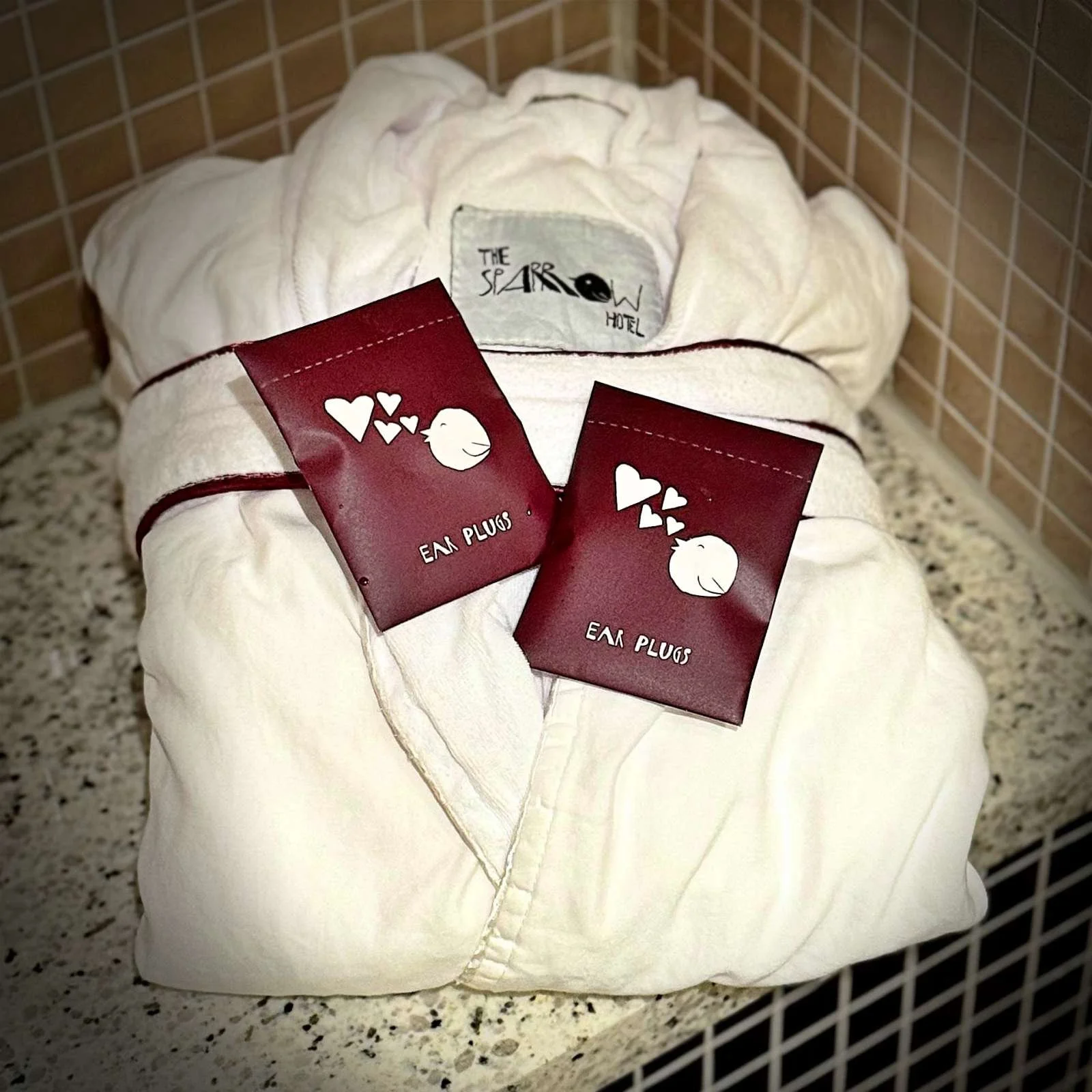 Ein zusammengelegter weißer Frottee-Bademantel mit dem Logo des Hotels im Kragen. Auf dem Mantel liegen zwei rote Papierpackungen mit der weißen Aufschrift „EAR PLUGS“.