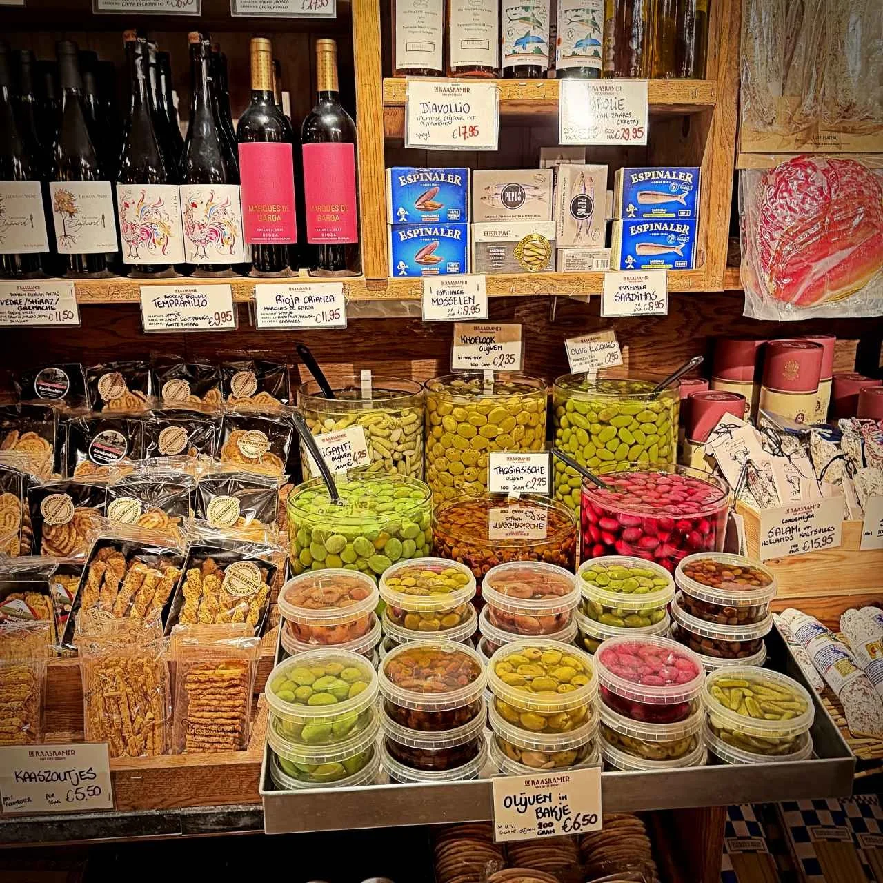 Im Shop De Kaaskamer in Amsterdam stehen verschiedene Oliven in großen Gläsern und Bechern. Darüber befinden sich Regale mit Weinflaschen, Fischkonserven und Kisten mit herzhaftem Gebäck.