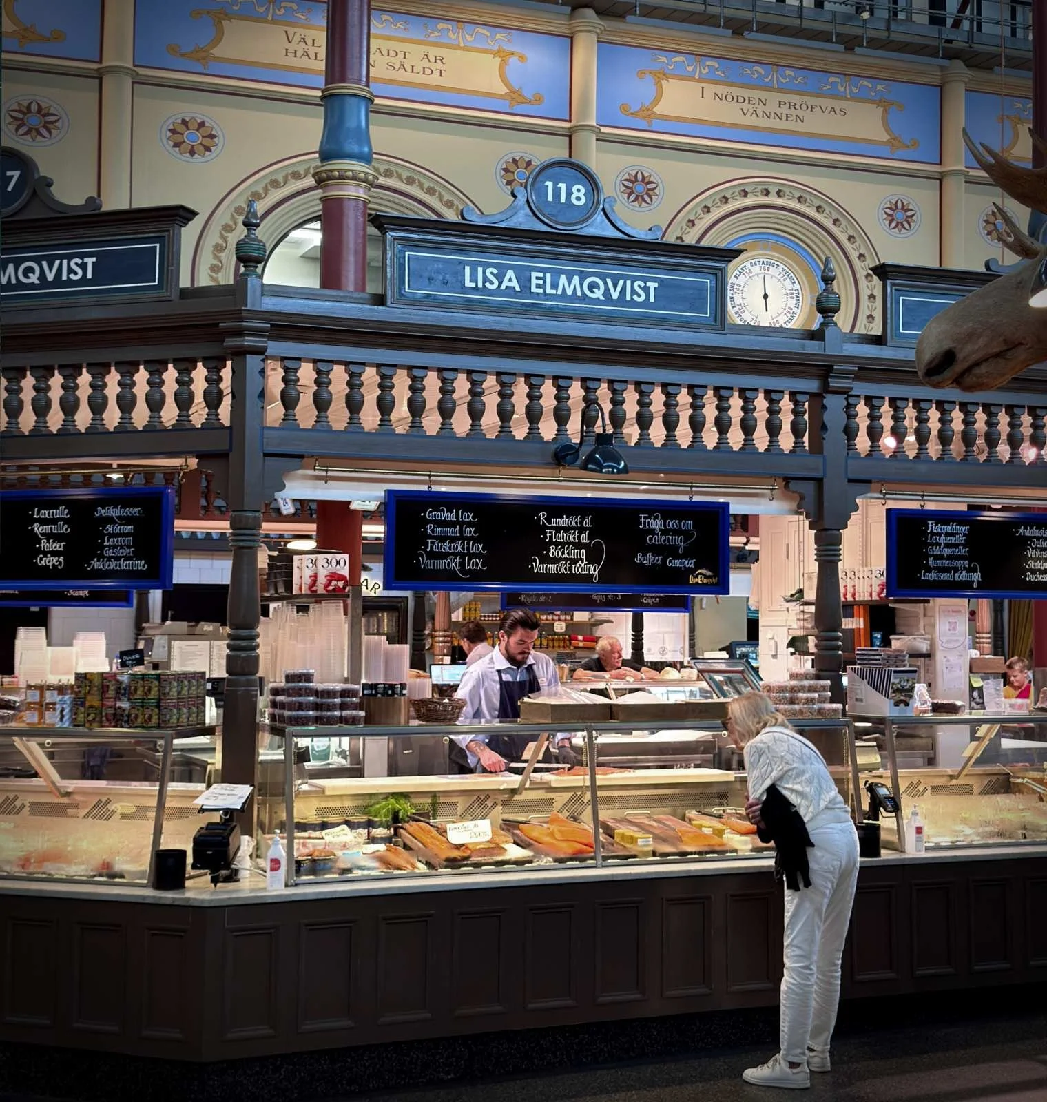 Eine Kundin betrachtet die Auslage am Stand von Lisa Elmqvist in der Östermalms Saluhall. Hinter der Glastheke mit verschiedenen Lachsspezialitäten bereitet ein Mitarbeiter Bestellungen vor.