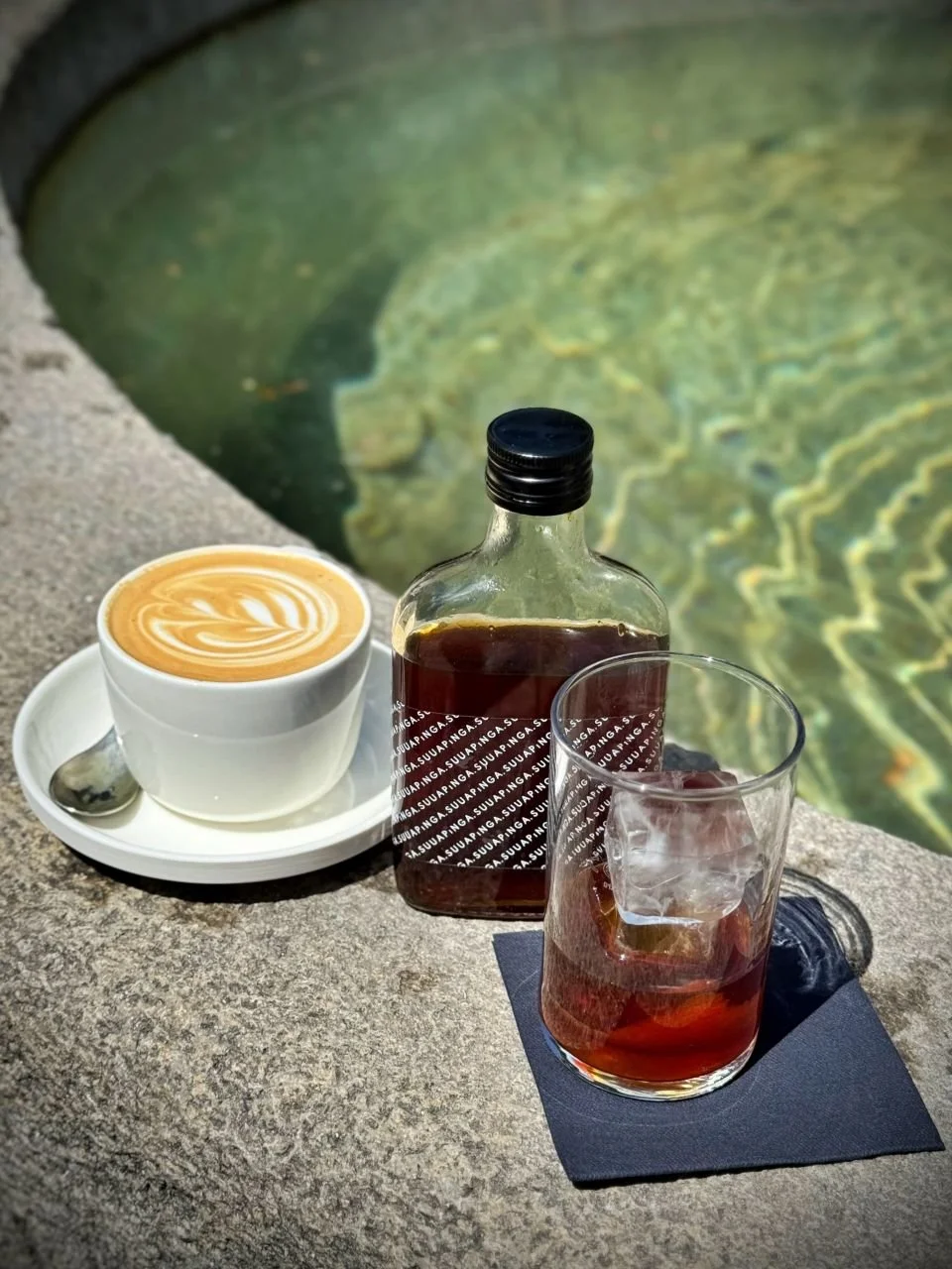 Ein Cold Brew mit Eiswürfeln, eine kleine Glasflasche und ein Cappuccino mit Latte Art stehen auf dem Rand eines Steinbrunnens am Wiener Platz in München. Die Getränke stammen vom Café Suuapinga.