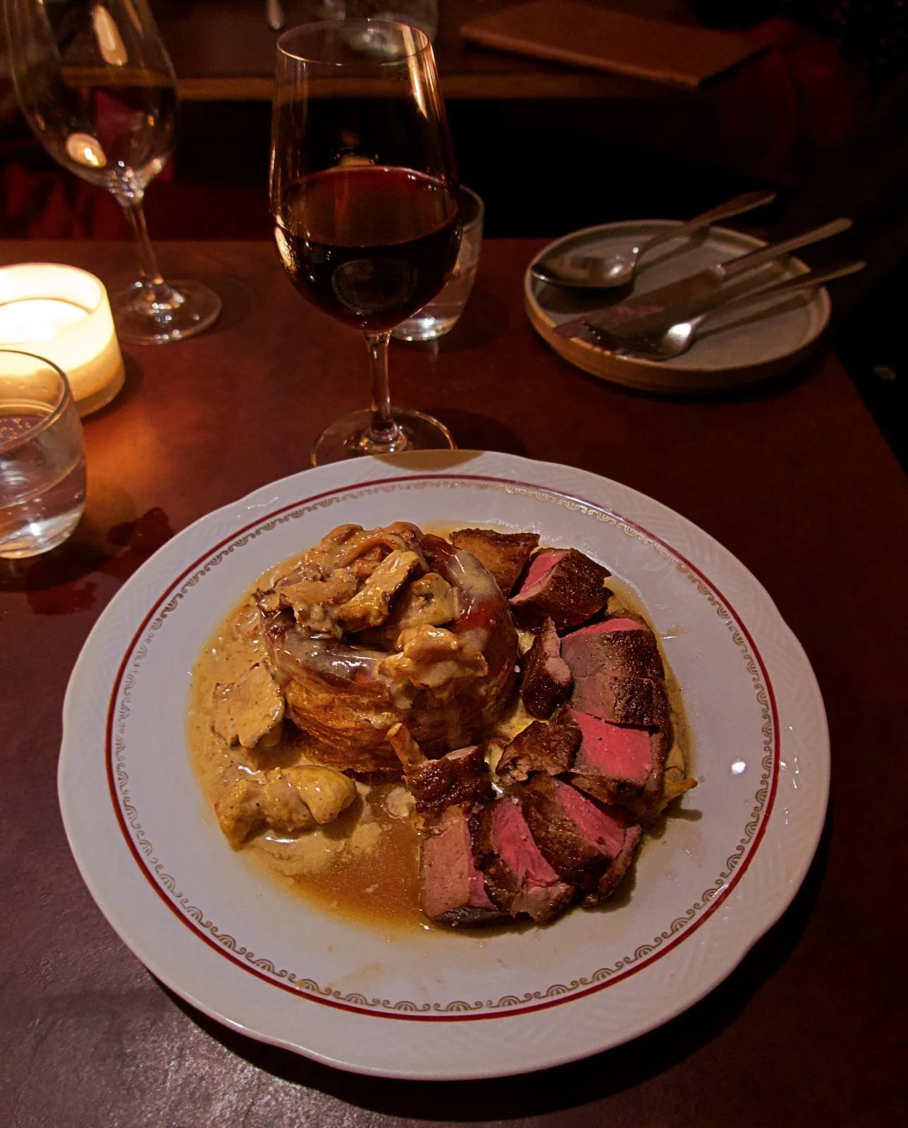 Königinnen-Pastete mit Pilzen und Sauce, serviert neben rosa gebratener Entenbrust auf einem Porzellanteller im Restaurant Främmat in Stockholm. Dazu wird ein Glas Rotwein bei warmem Kerzenschein gereicht.