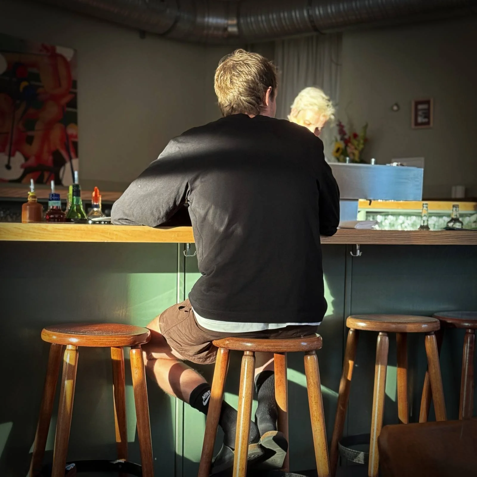 Rear view of a man sitting on a wooden stool at the green bar counter of Restaurant Trio. Bottles stand on the counter as sunlight shines in from the side.