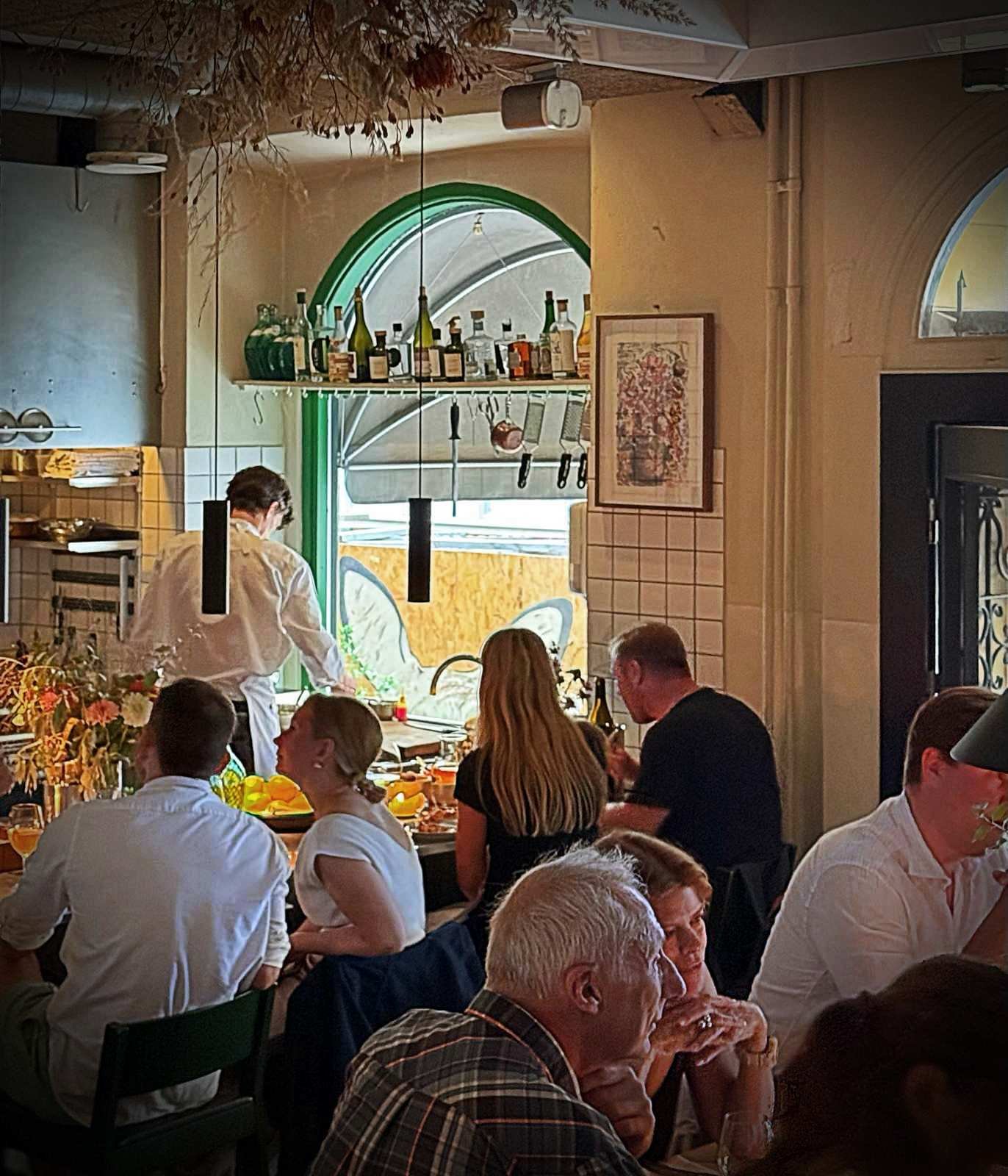 Gastraum eines Restaurants mit besetzten Tischen im Vordergrund. Im Hintergrund steht ein Koch hinter einem Tresen vor einer gefliesten Wand mit Wandregal.