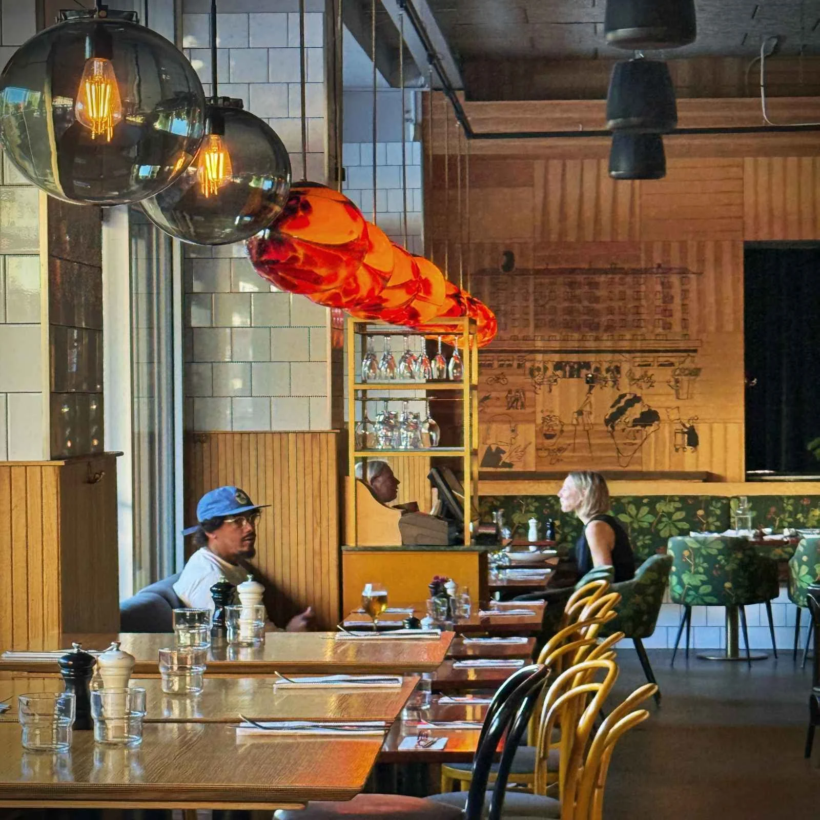 The dining area of a restaurant featuring set wooden tables, yellow and black chairs, and orange-red glass globe pendant lights. Several people are seated at tables, with illustrated wooden paneling visible in the background.