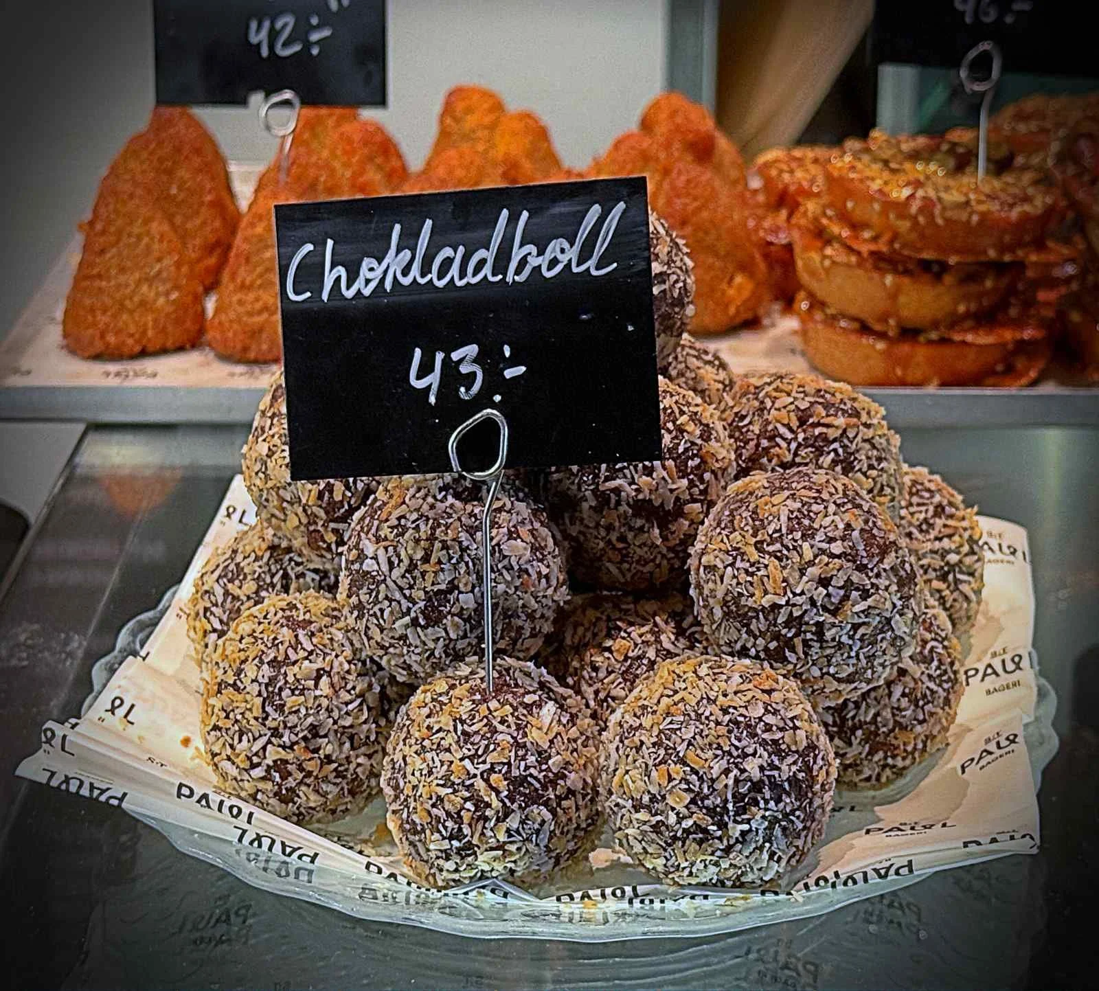 A stack of Swedish chocolate balls (Chokladbollar) coated in coconut flakes on a glass plate. A small black price tag reading "Chokladboll 43:-" is placed in the center.