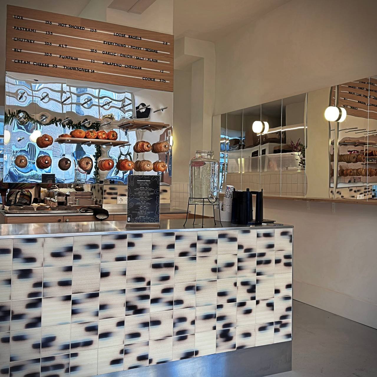 Theke mit hängenden Bagels im Café Breadwinner Bagels im Amsterdamer Museumkwartier, Holzregale, Preistafel an der Wand, Glaswasserspender und geflieste Front im Innenraum.