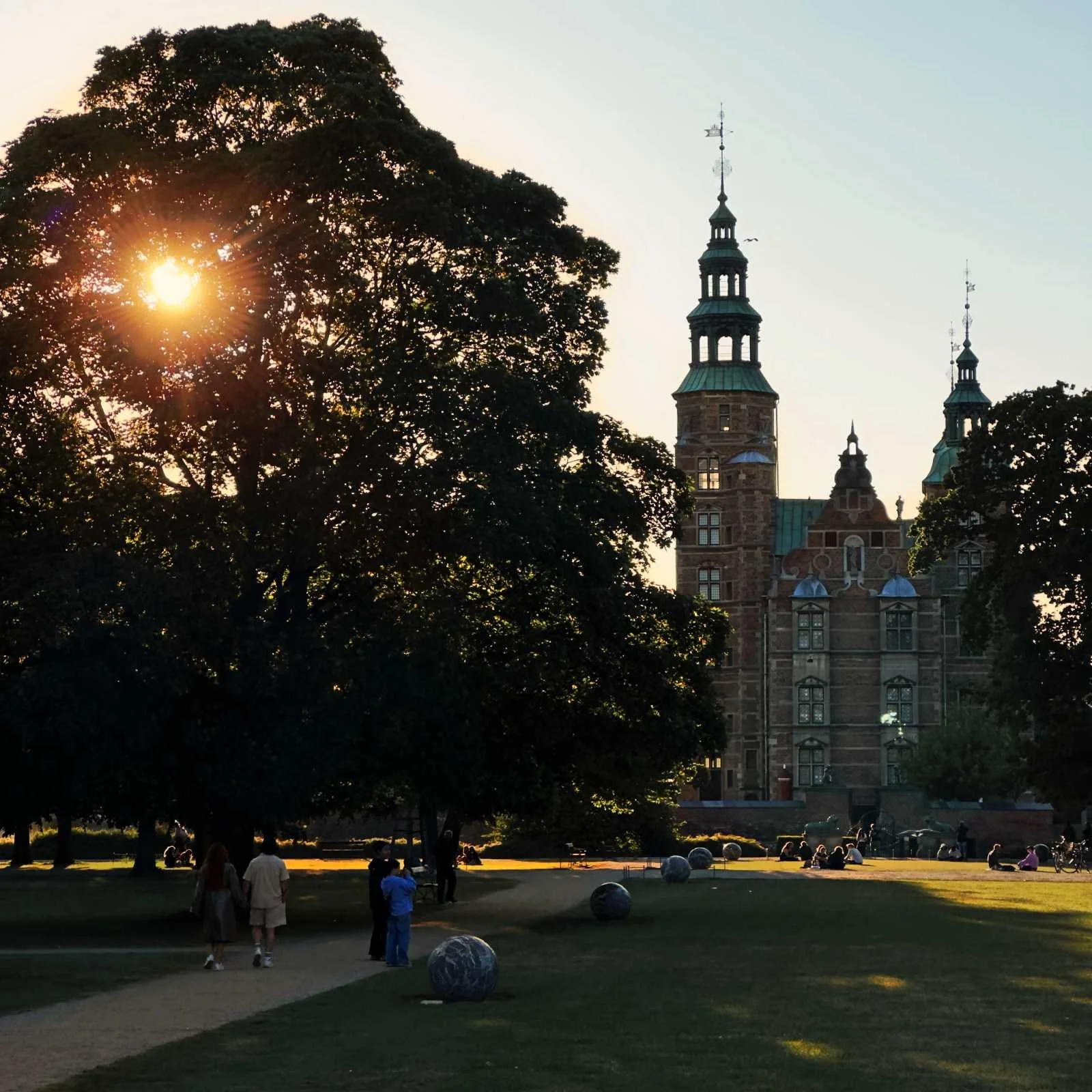 View through Kongens Have in Copenhagen towards Rosenborg Castle with brick towers and copper roofs. Lawn with footpath, trees, stone sculptures and people relaxing in the park.