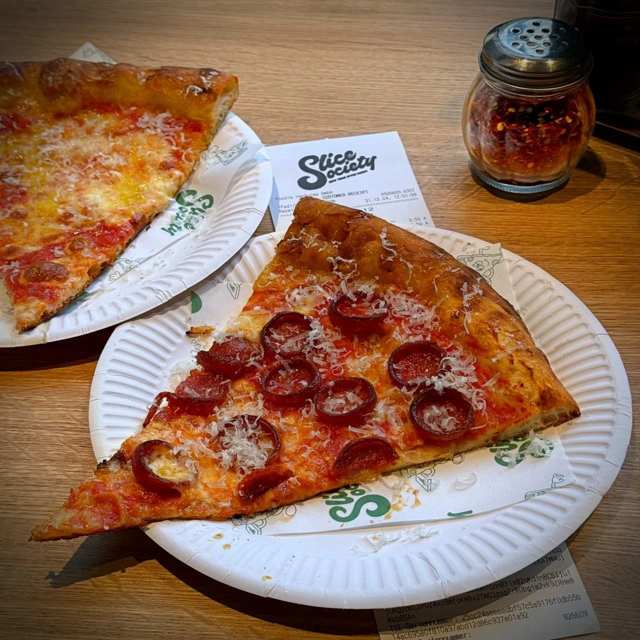 Two large slices of cheese and salami pizza on white paper plates, served at Slice Society in Berlin with receipt and chili flakes shaker in the background.
