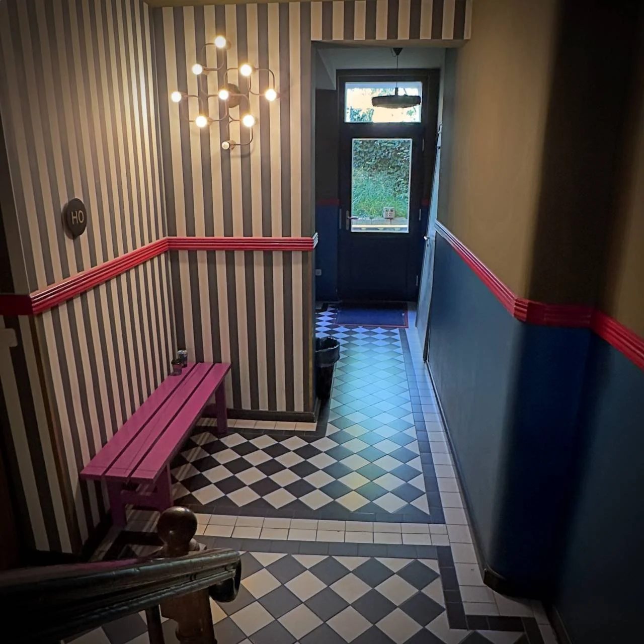A hallway at Brilliant Apartments in Berlin featuring striped wallpaper and a black and white checkered tile floor. The interior includes a purple bench and a modern wall lamp on a blue wall with red trim.