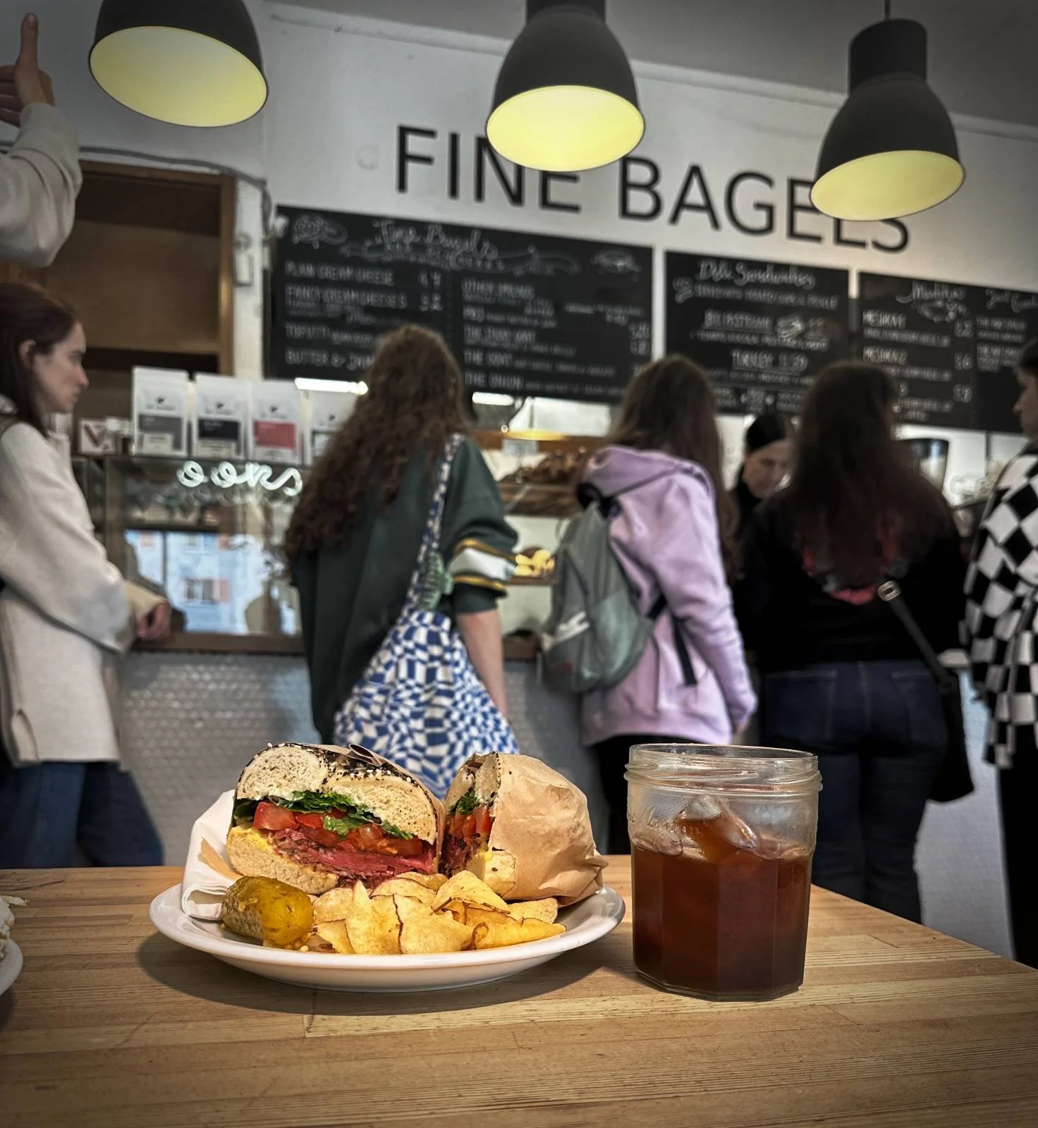 Ein belegter Bagel mit Chips und einer Gewürzgurke auf einem Teller bei Shakespeare and Sons in Berlin. Daneben steht ein Eiskaffee im Glas vor der Theke mit dem Schriftzug „Fine Bagels“.