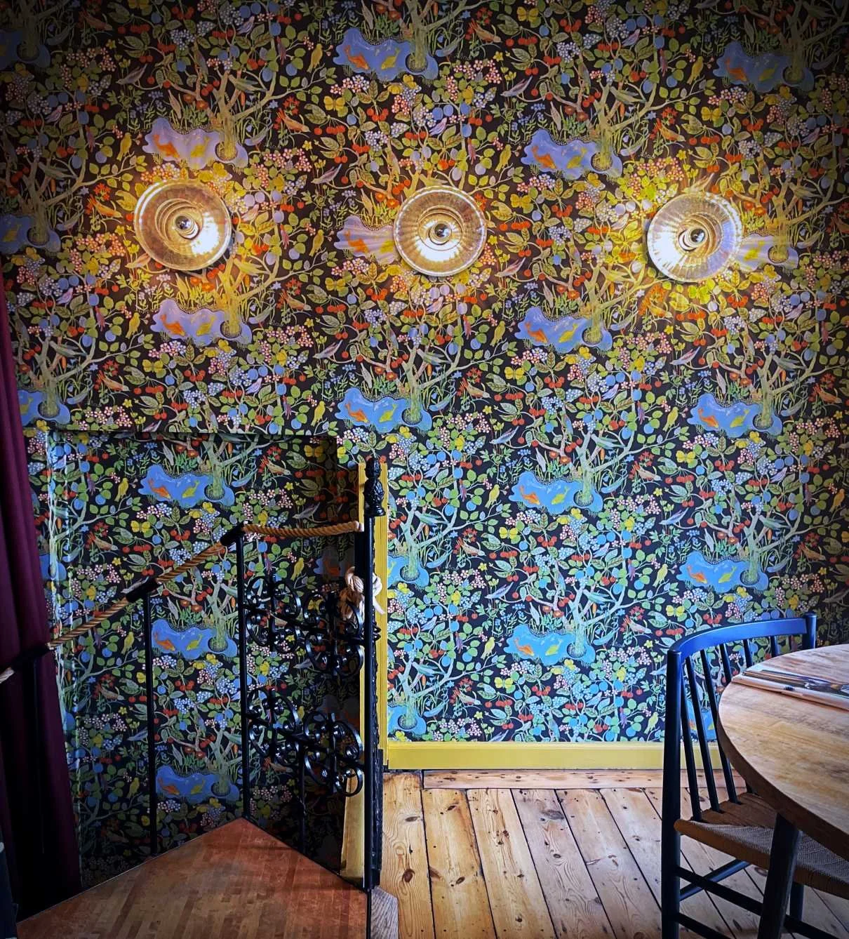 Die Wand im Kopenhagener Restaurant Selma ist mit der bunten Tapete „Paradiset“ von Josef Frank tapeziert. Davor befinden sich eine schwarze Wendeltreppe, drei runde Wandleuchten und ein Holztisch.
