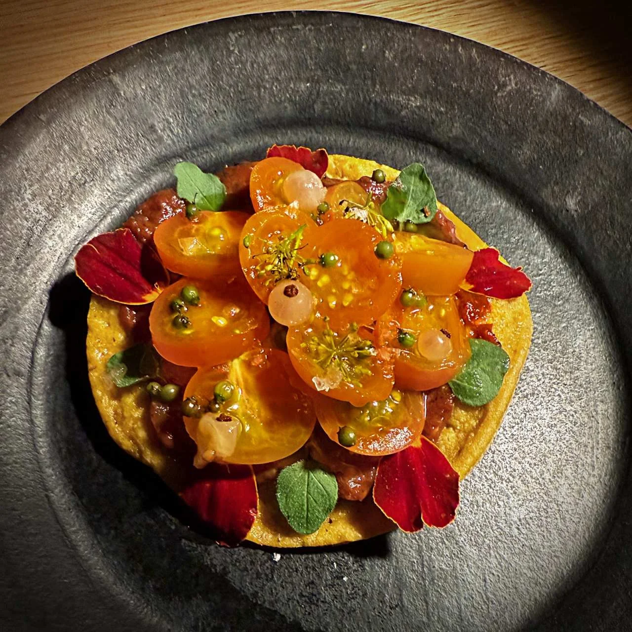 Tostada mit gelben Tomaten, weißen Johannisbeeren und roten Blütenblättern serviert auf einem dunklen Teller im Restaurant Sanchez in Kopenhagen.
