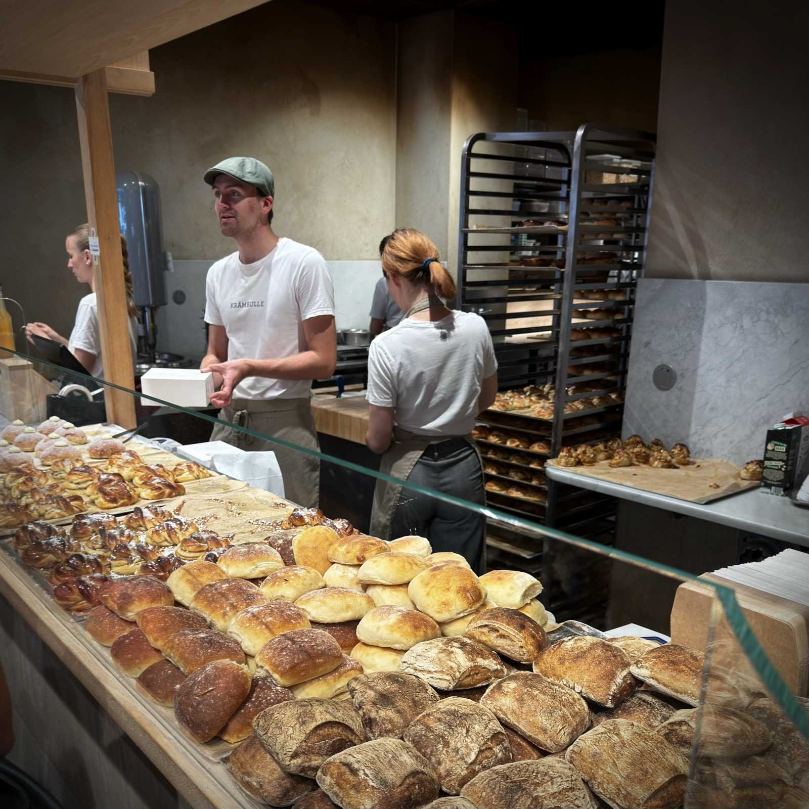 Eine lange Glasvitrine gefüllt mit verschiedenen Brötchen, Zimtschnecken und gefülltem Gebäck. Dahinter stehen Mitarbeiter der Bäckerei in weißer Kleidung bei der Arbeit, im Hintergrund ist ein hoher Stufenwagen mit Backblechen zu sehen.