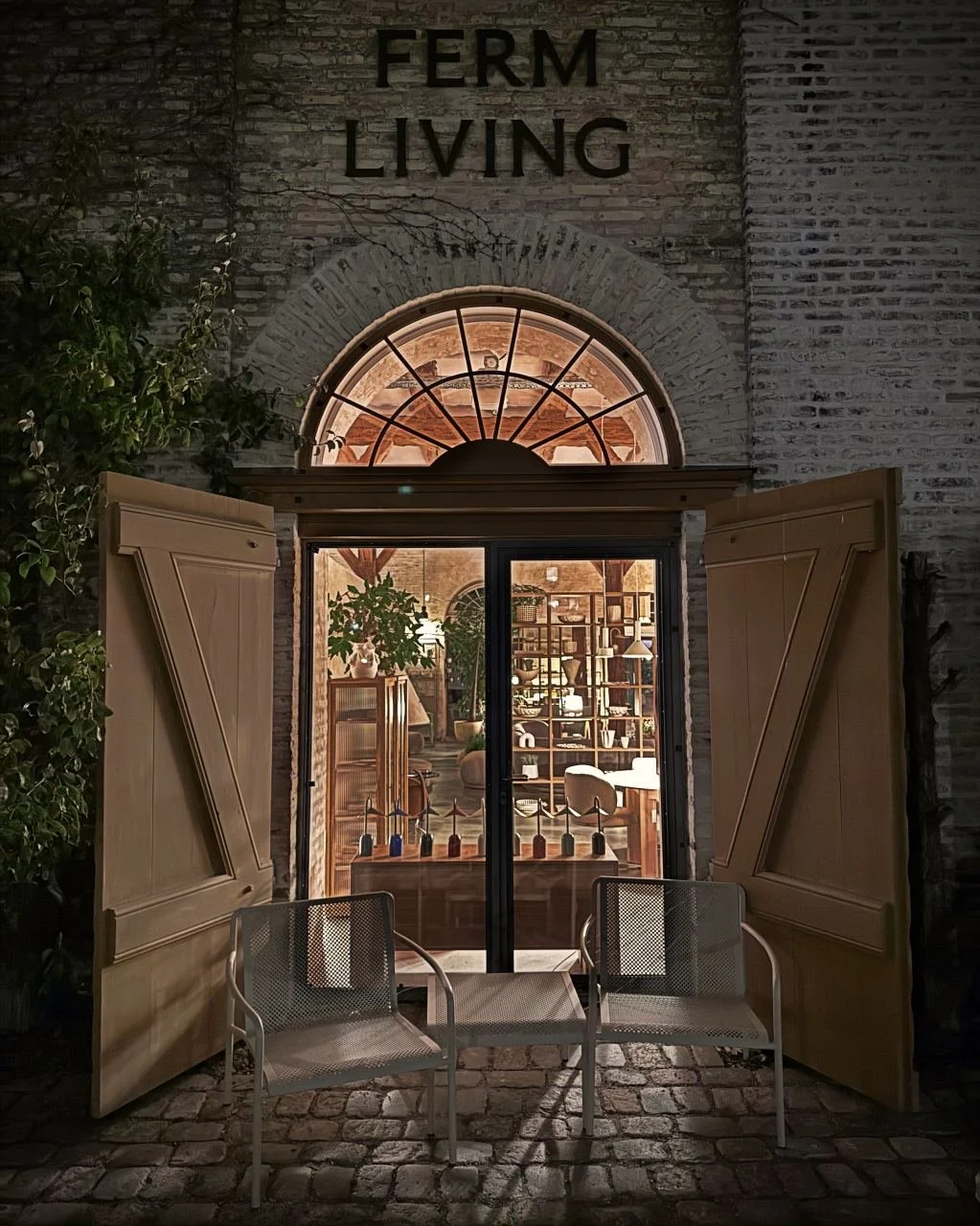 Open wooden doors reveal the Ferm Living store on Paper Island in Copenhagen, warmly lit at night with interior design shelves and two white metal chairs in front.