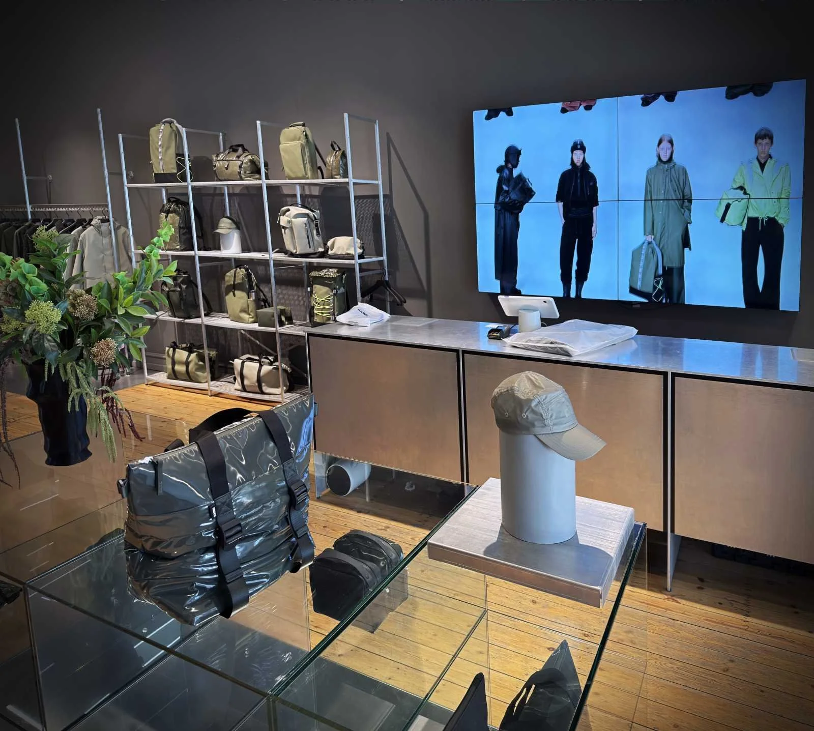 Präsentation von Taschen und einer Kappe auf Glastischen und Regalen im Rains Store vor einer Videowand.