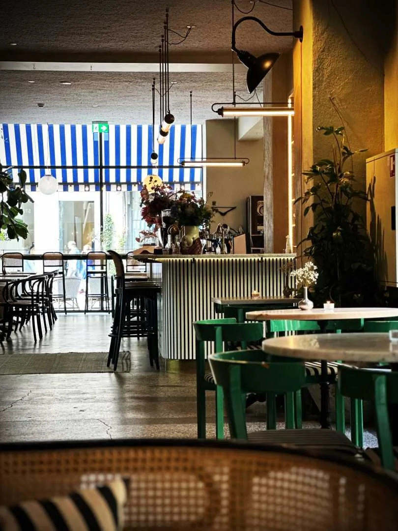View into the lobby and bar area at the Coco Hotel in Copenhagen. The room is furnished with green wooden chairs, round tables, and a counter with hanging lamps. A blue and white striped awning is visible through the window front.