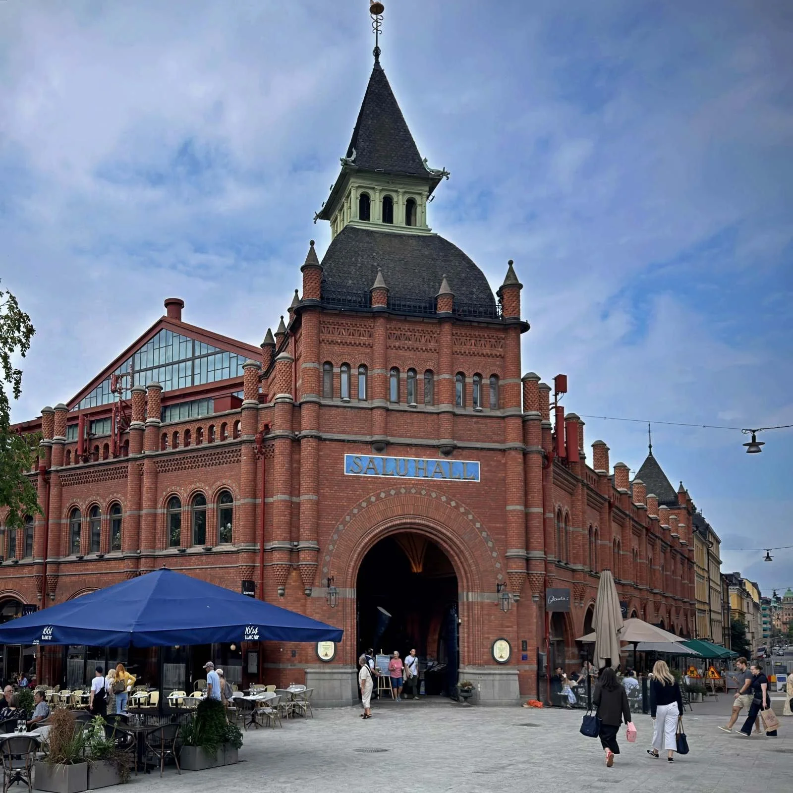 Die markante rote Backsteinfassade der historischen Östermalms Saluhall in Stockholm mit ihrem charakteristischen Eckturm. Vor dem Haupteingang am Östermalmstorg befinden sich Außenterrassen von Cafés unter blauen Sonnenschirmen.