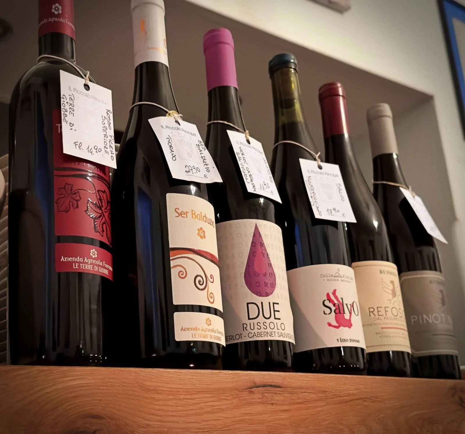 Wine bottles with handwritten price tags lined up on a wooden shelf at Il Piccolo Principe restaurant in Munich.