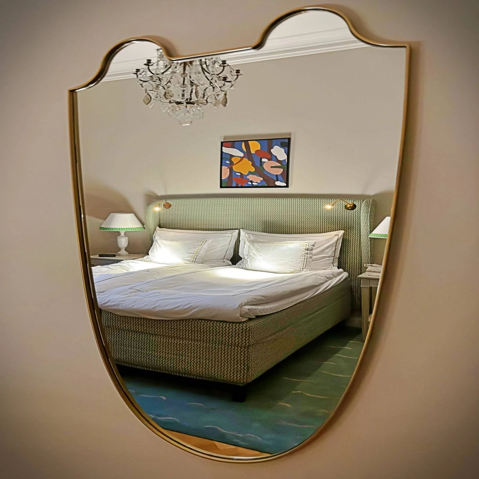 Spiegelung eines Hotelzimmers in einem an der Wand hängenden Spiegel mit geschwungenem Goldrahmen. Im Spiegel sichtbar sind ein Doppelbett mit grün gemustertem Kopfteil und weißer Bettwäsche