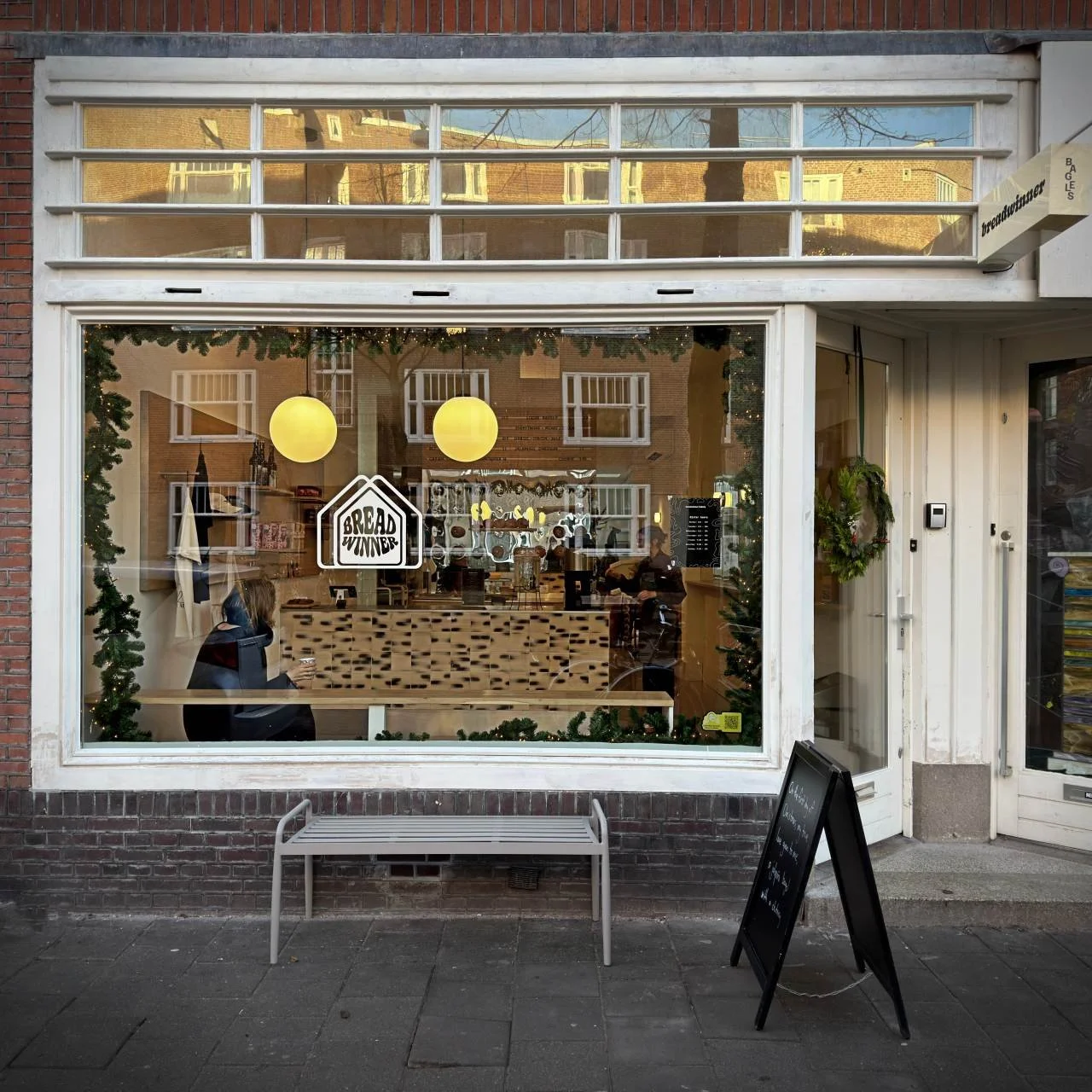 Exterior view of Breadwinner Bagels café in Amsterdam’s Museum Quarter with large front window, logo on the glass, view of counter and guests inside, bench in front.