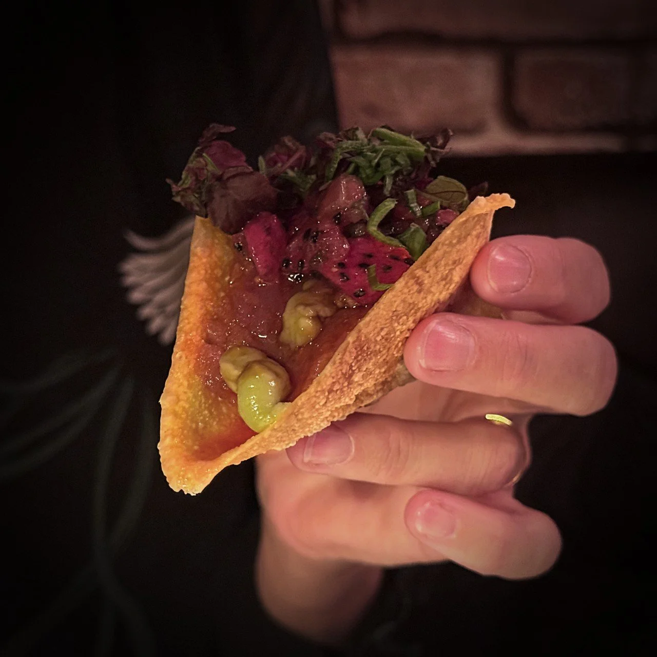 Nahaufnahme eines knusprigen Tacos in der Hand, gefüllt mit rohem Fisch, Edamame, Drachenfrucht, Blattsalat und asiatischer Sauce, vor dunklem Hintergrund.