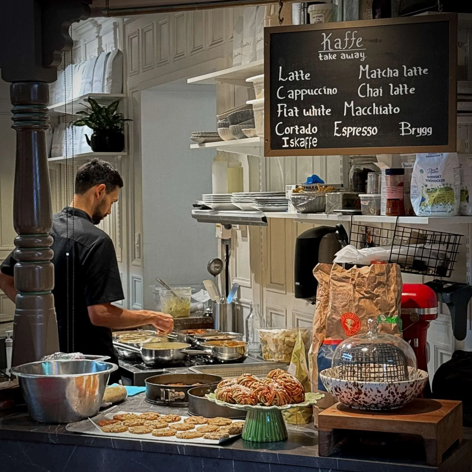 A staff member works behind the counter in a cafe inside Östermalms Saluhall. Fresh cinnamon buns and cookies are displayed on the counter, with a hand-written chalkboard menu for coffee hanging above.