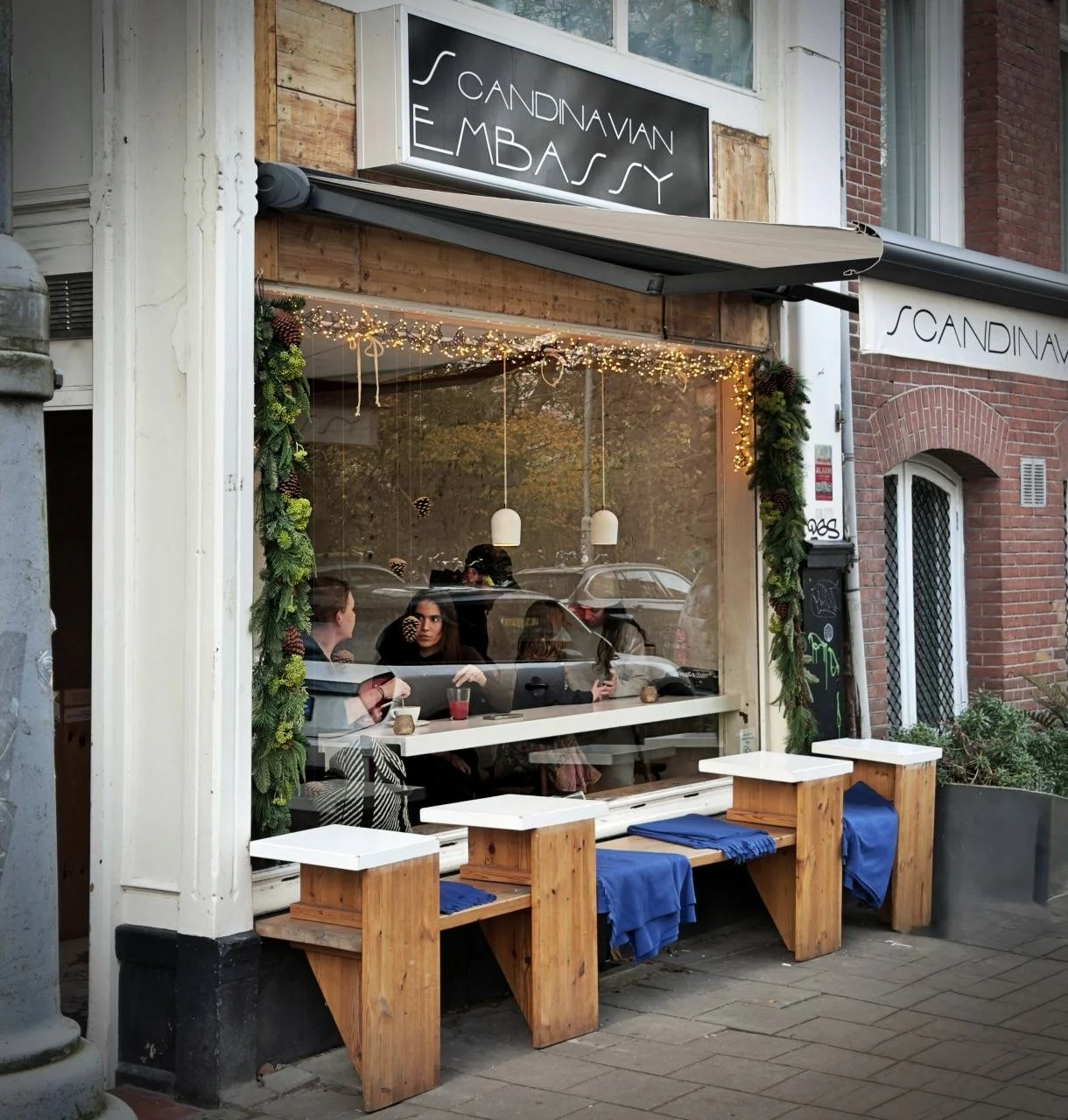 Außenansicht des Cafés Scandinavian Embassy in Amsterdam mit großer Fensterfront, Sitzplätzen an der Fassade, Holzbänken mit blauen Kissen und Gästen im Innenraum hinter der Scheibe.
