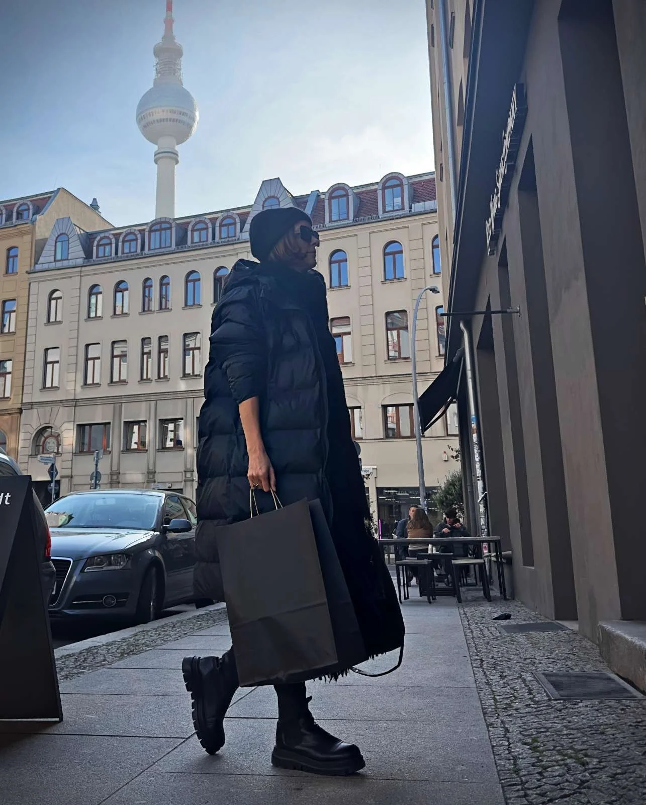 Eine Frau in einem langen schwarzen Daunenmantel geht mit einer Einkaufstüte über einen Gehweg, im Hintergrund ist der Berliner Fernsehturm zu sehen.