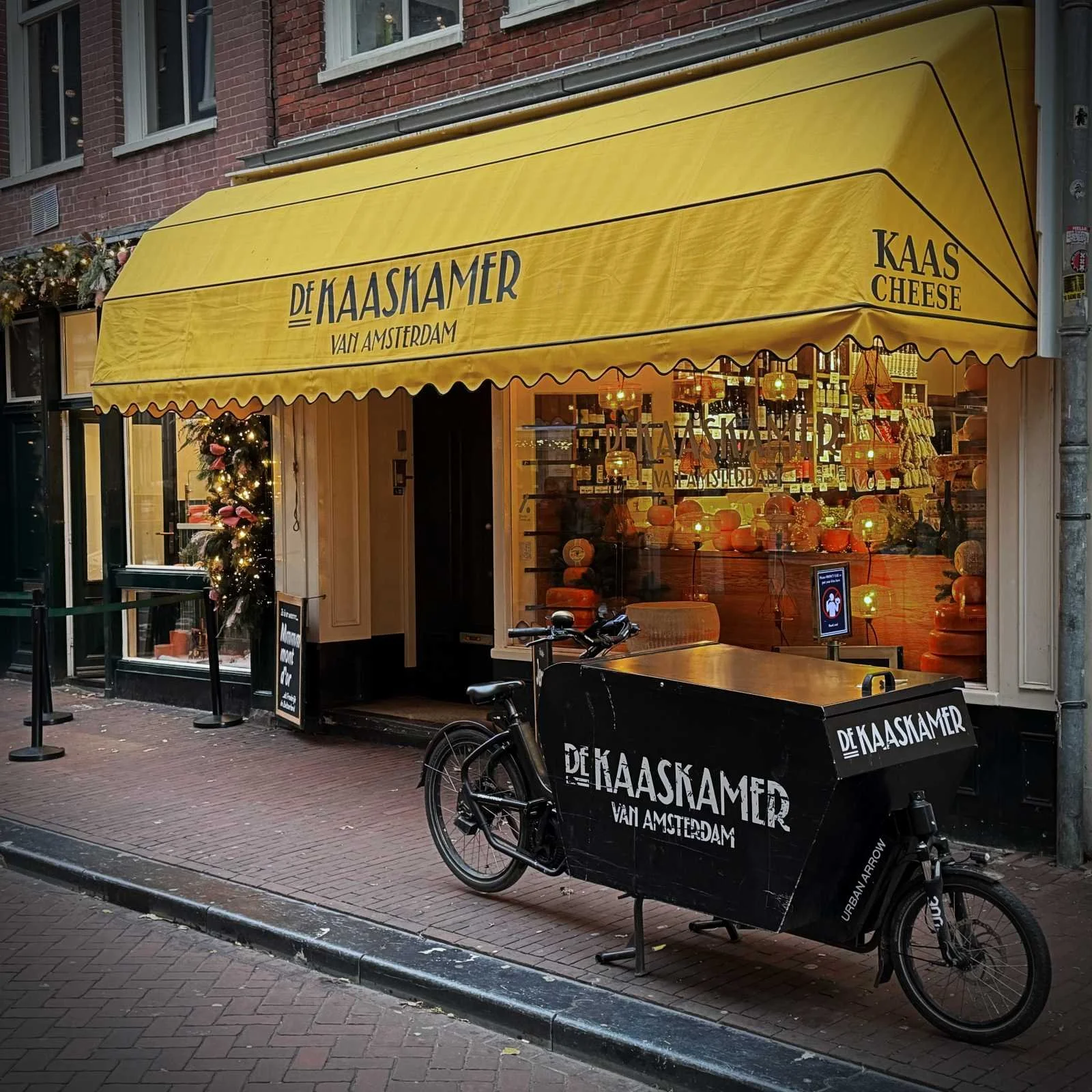 A black cargo bike is parked in front of De Kaaskamer shop in Amsterdam. A yellow awning with the shop's name hangs over the window, which displays numerous cheese wheels and interior lights.