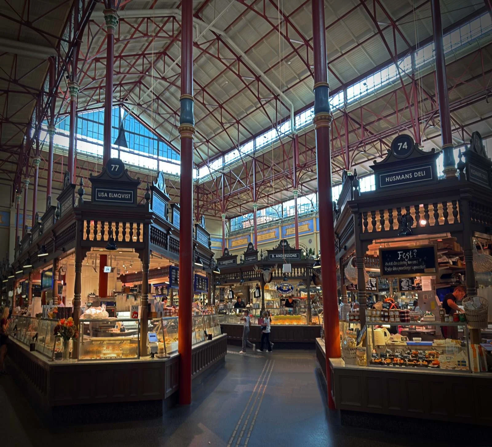 Die Innenaufnahme zeigt die großzügigen Gänge der Östermalms Saluhall in Stockholm. Unter der ikonischen roten Dachkonstruktion aus Metall präsentieren sich traditionsreiche Marktstände wie „Lisa Elmqvist“ und „Husmans Deli“.