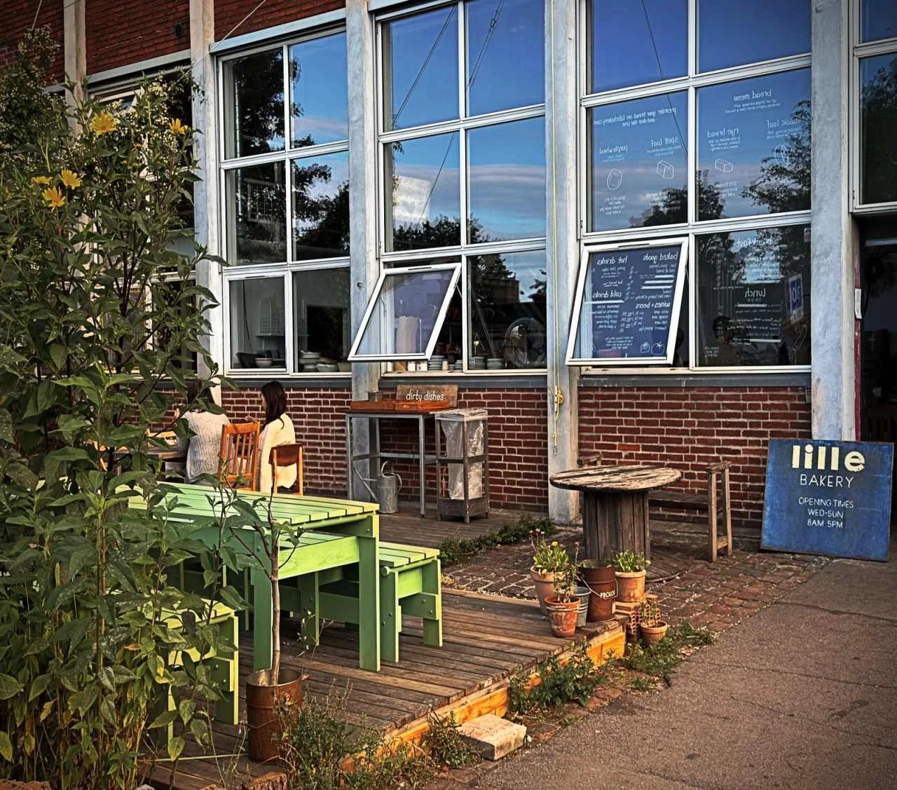 Außenbereich der Lille Bakery in Kopenhagen mit grünen Holztischen vor einer Backsteinfassade. Ein blaues Schild zeigt die Öffnungszeiten, daneben wachsen hohe gelbe Blumen.