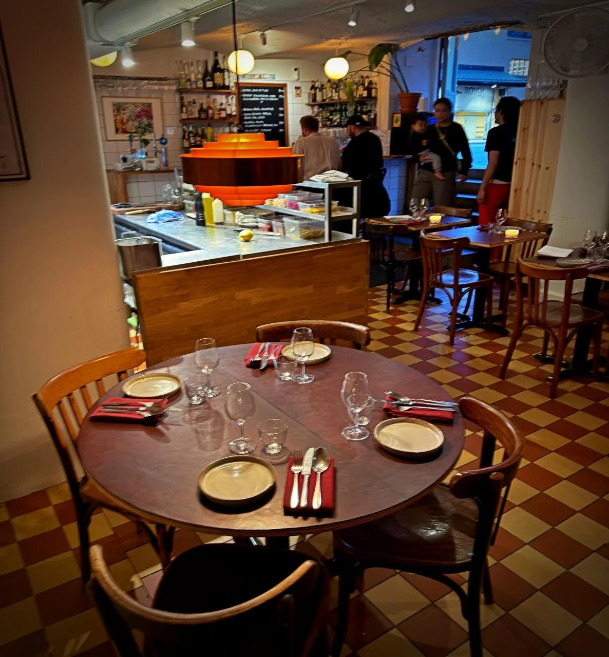 Die Aufnahme zeigt den einladenden Innenraum des Restaurants Främmat in Stockholm mit gedeckten Holztischen, einem klassischen Schachbrettboden und einer atmosphärischen orangefarbenen Lampe über dem Tresen.